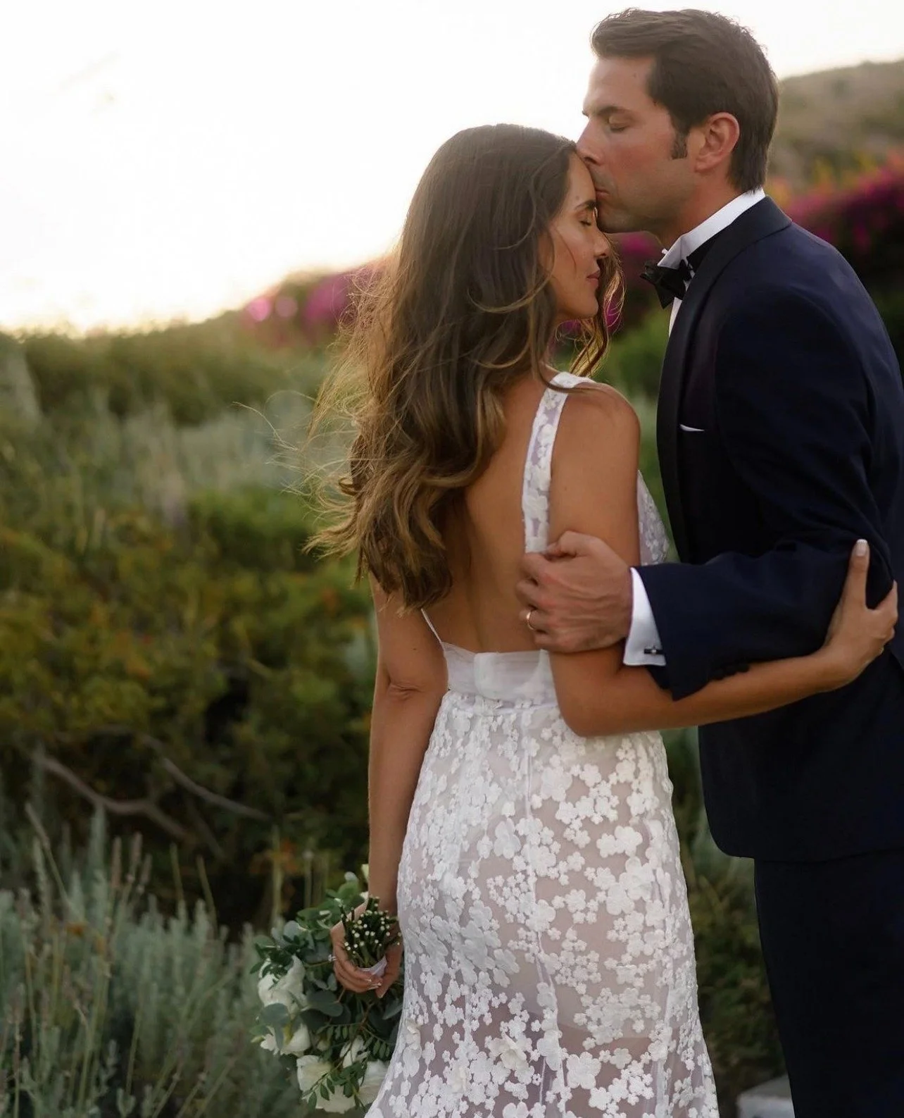 A bride in a white lace wedding dress holding a bouquet and a groom in a tuxedo sharing a kiss outdoors during sunset.