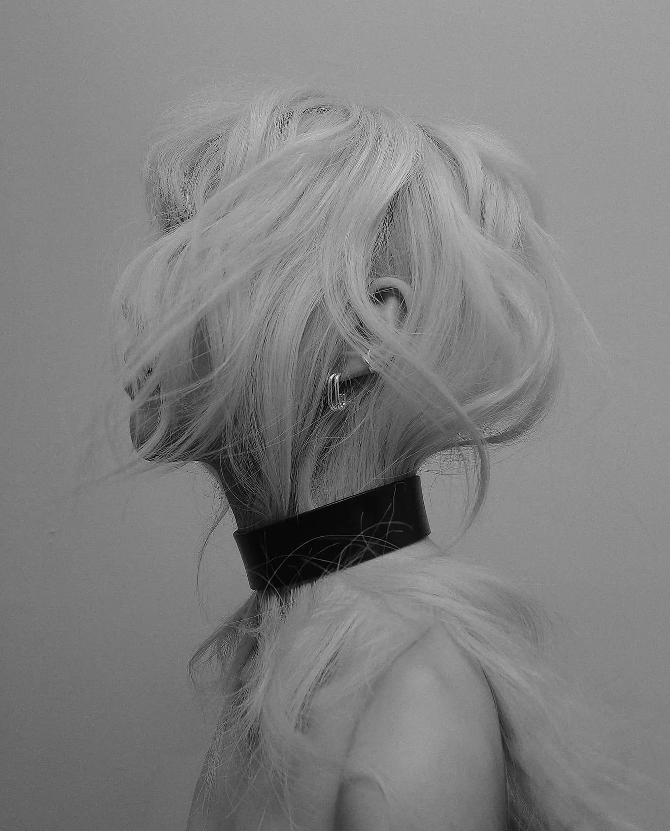 Black and white photo of a person's head with tousled platinum blonde hair, seen from the back, wearing large hoop earrings and a black choker.