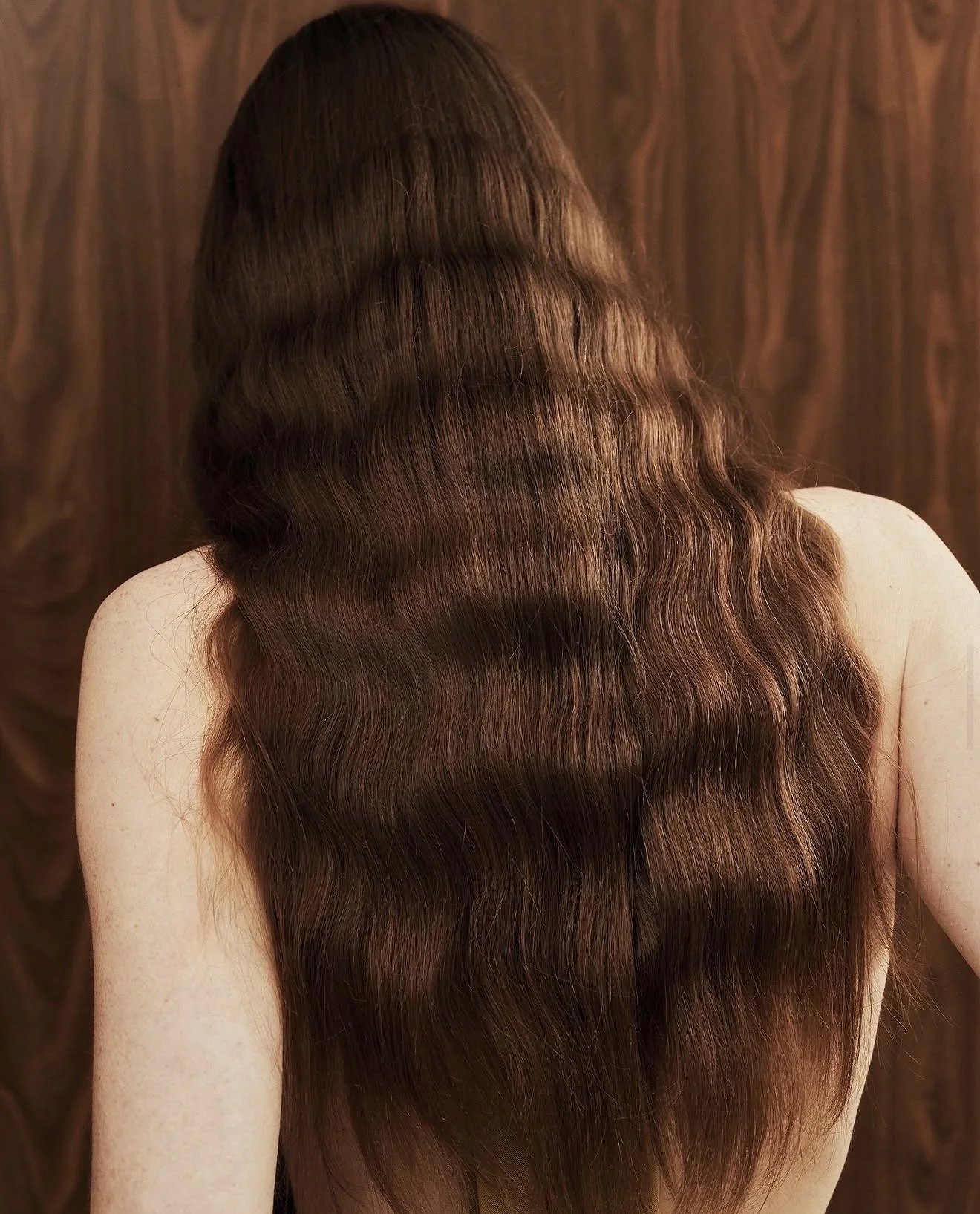 Back view of a woman with long, wavy, brown hair against a wooden background.