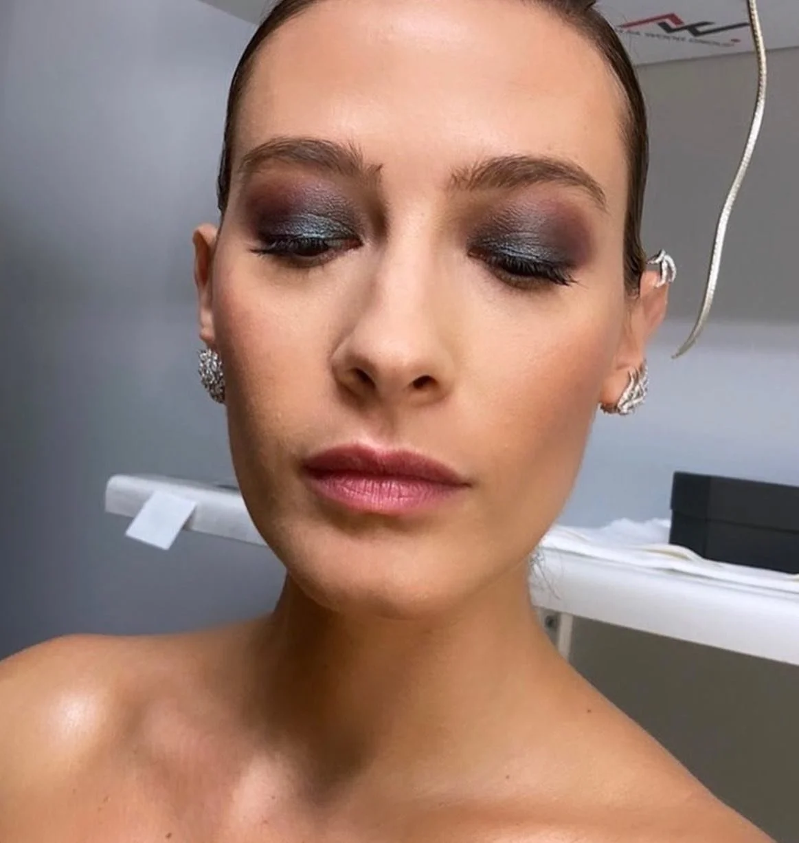 Close-up of a woman with makeup, closed eyes, and earrings, in a room with medical equipment in the background.