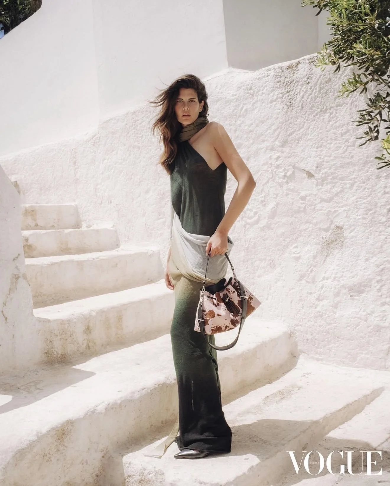 A woman standing on white steps made of plaster or concrete, with a white wall and some greenery nearby. She has long, dark hair and is wearing a black sleeveless top, dark green pants, and is carrying a bag with a giraffe print.