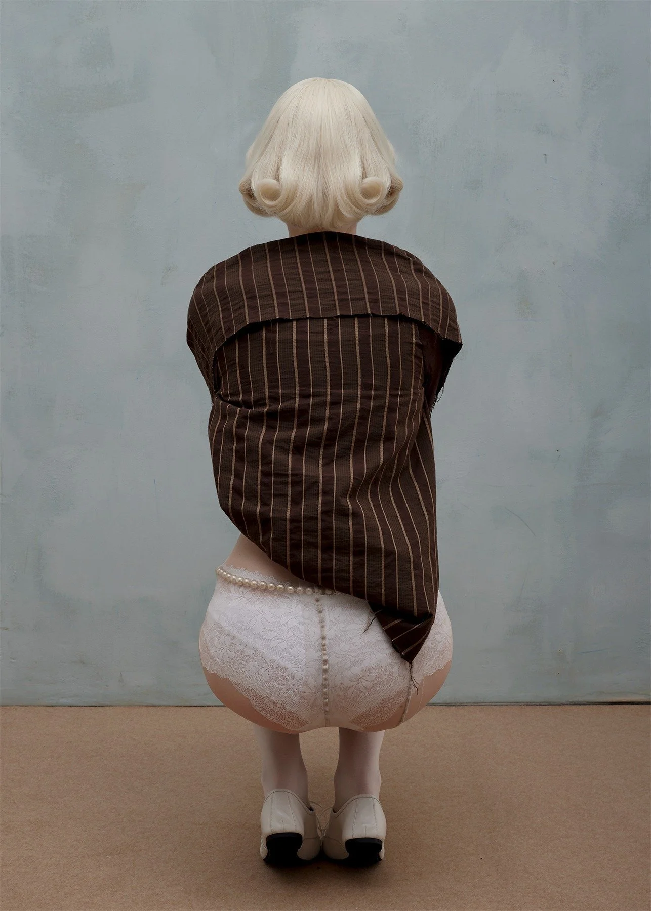 Back view of a person with platinum blonde hair styled in soft curls, wearing a brown pinstripe shirt, white lace underwear, white tights, and white shoes, standing against a plain light blue wall.