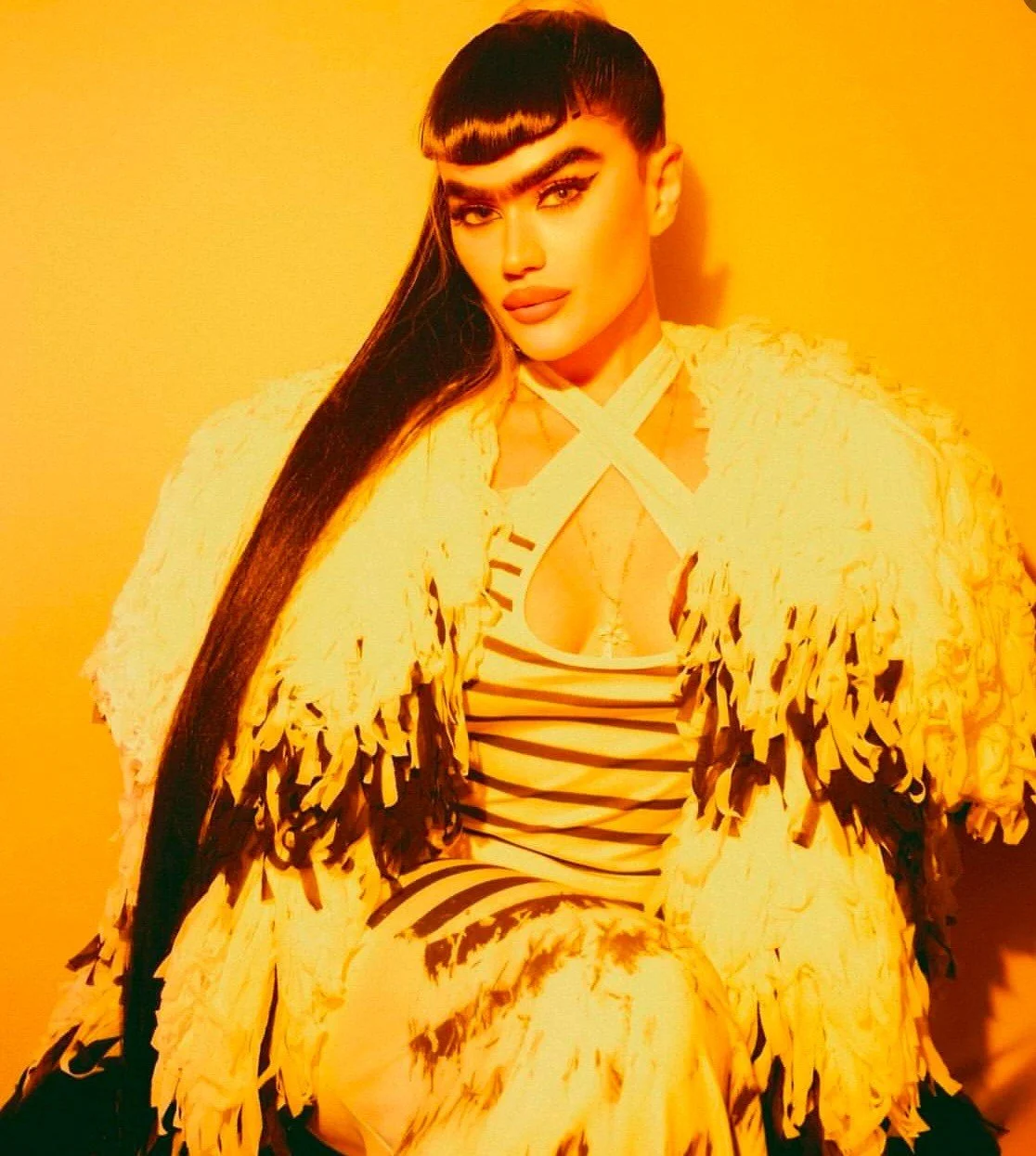 A woman with long dark hair, bold makeup, and a confident expression, sitting against a yellow background, wearing a fringed jacket and black and white striped top.