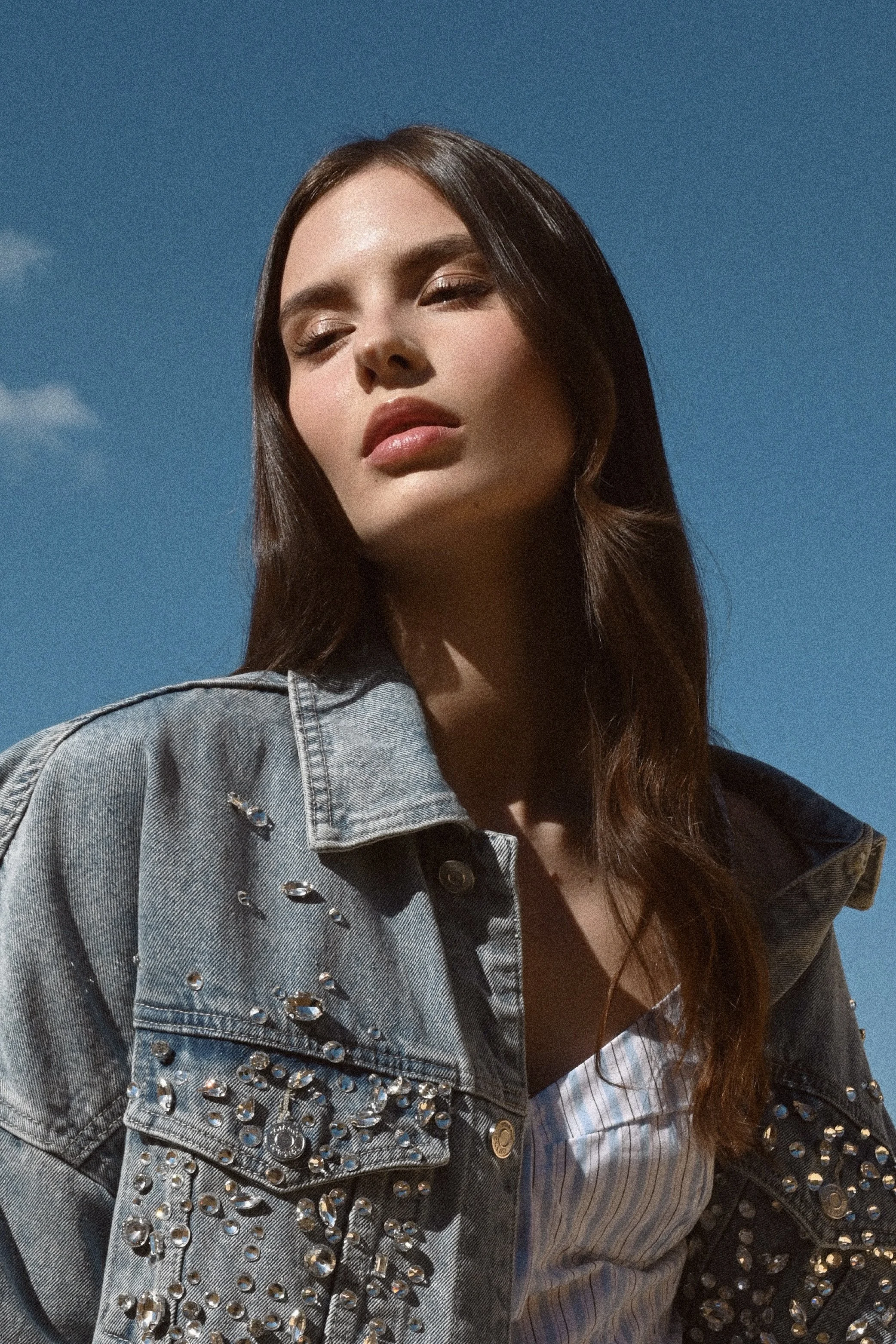 A young woman with long brown hair, wearing a denim jacket with rhinestones and a striped shirt, is outdoors against a clear blue sky.