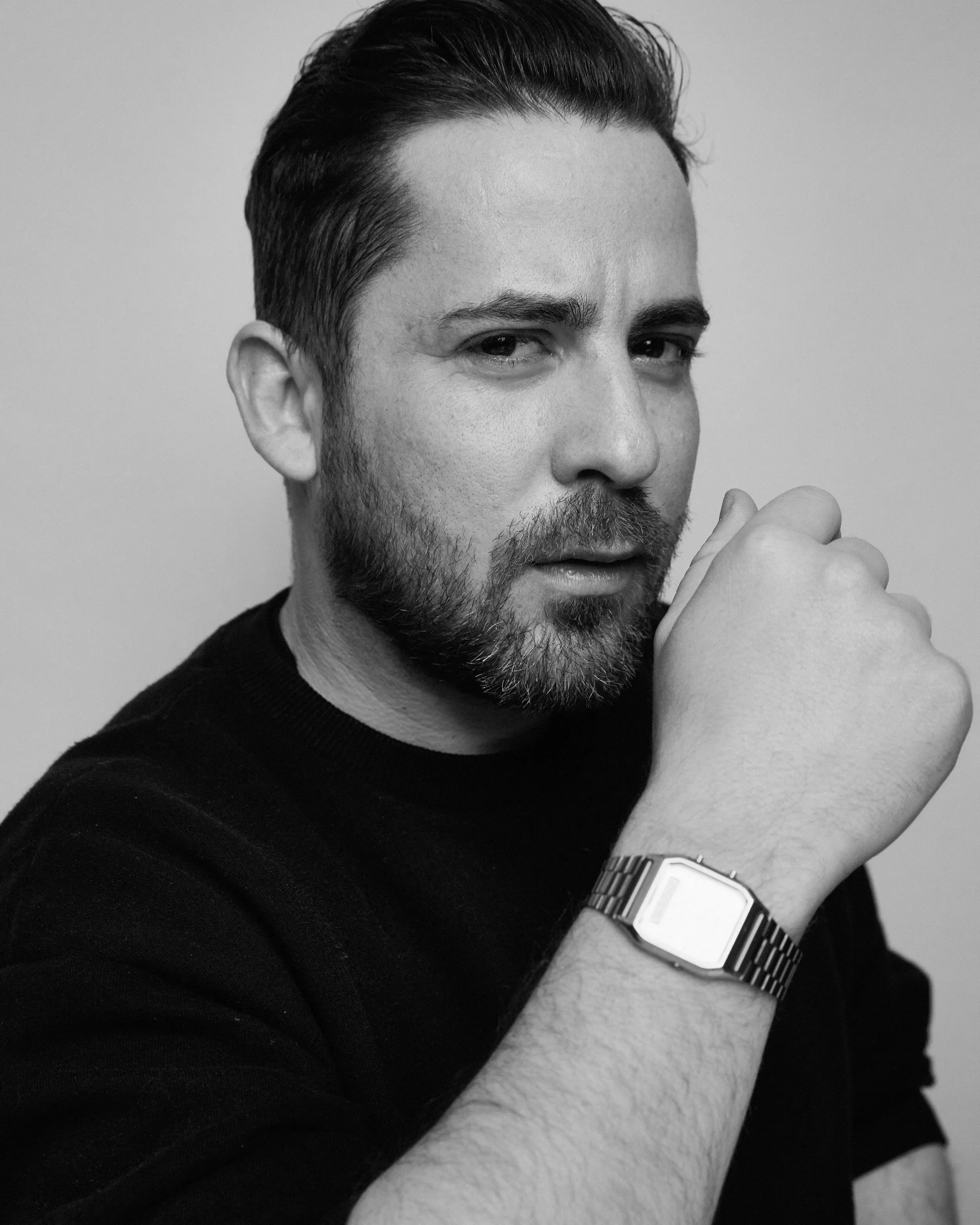 Black and white close-up photo of a man with dark hair and beard, wearing a watch and a black shirt, resting his chin on his hand, with a serious expression.