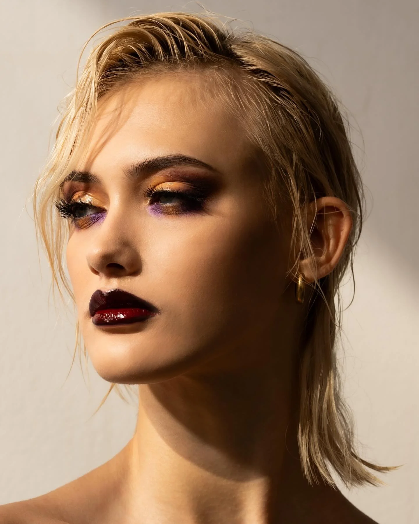 Close-up of a woman with short, wet blond hair, bold eye makeup featuring gold, purple, and dark shades, and deep red lipstick. She is wearing a gold hoop earring.