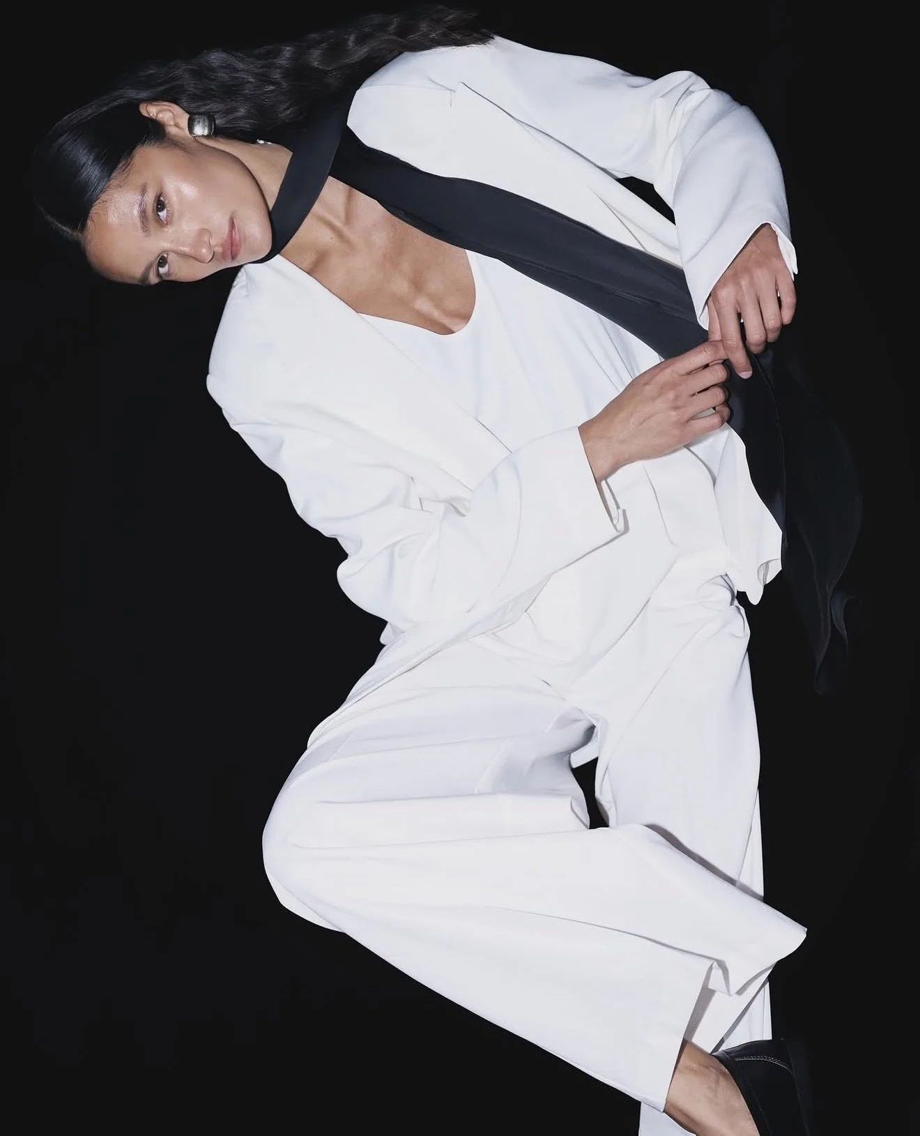 A woman with long dark hair wearing a white suit with black accents, lying against a black background.