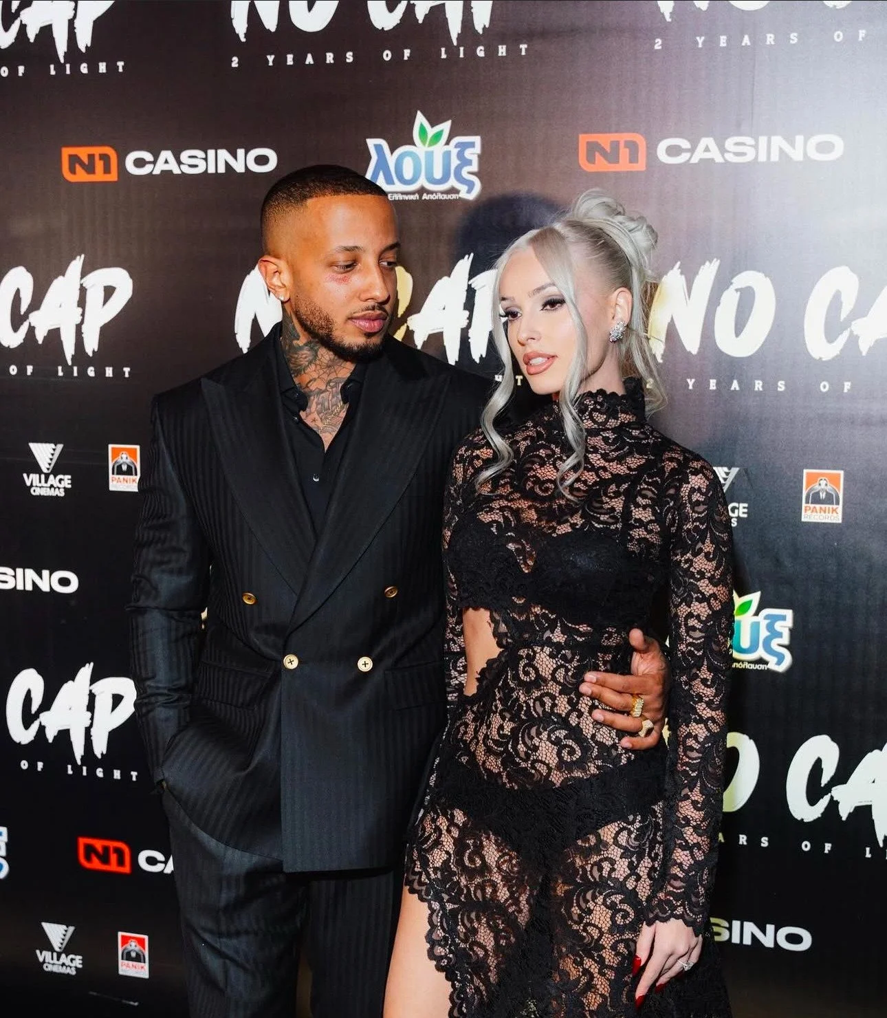 A man and woman posing together at a red carpet event, with a backdrop featuring various logos and text. The man is wearing a black pinstripe suit and has a tattooed neck, while the woman is in a black lace dress with silver hair styled in loose curl