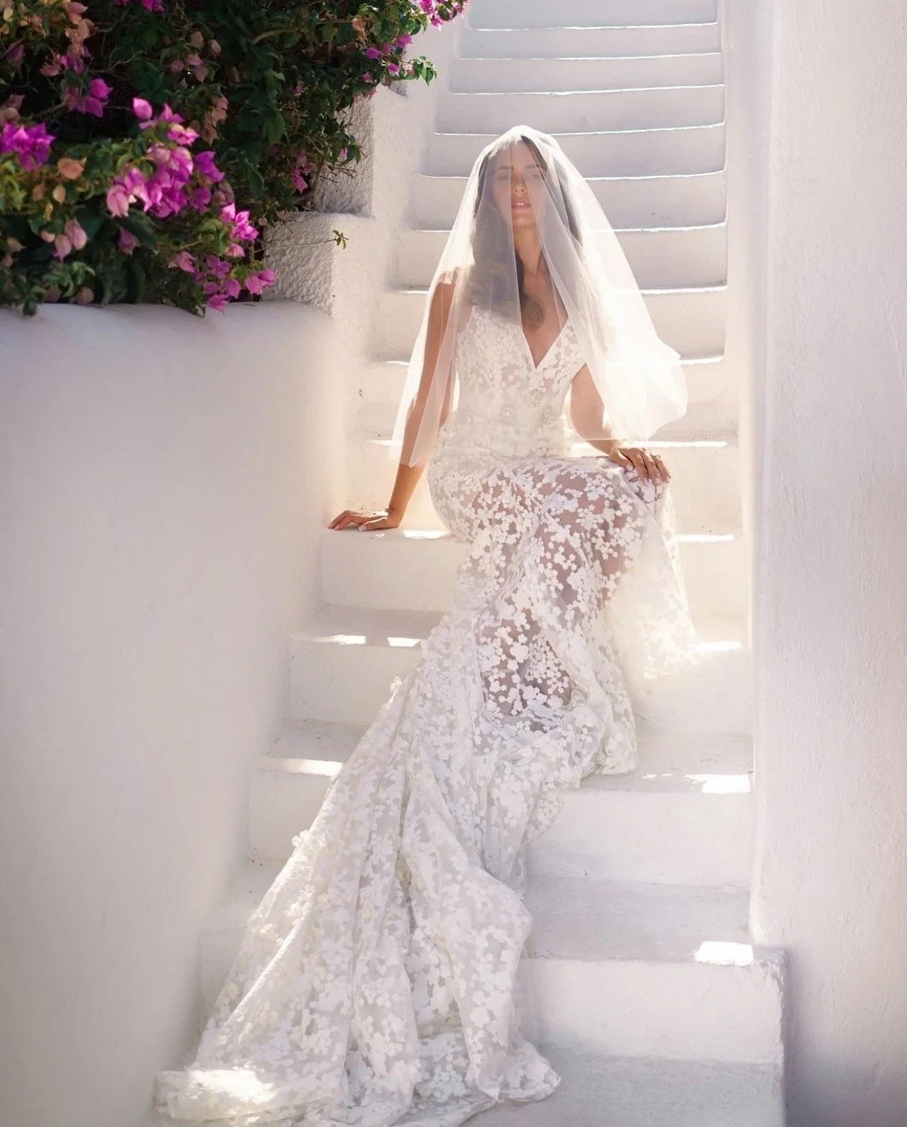 A woman wearing a lace wedding dress and veil sits on white stairs in front of a white wall, next to pink and purple flowers.
