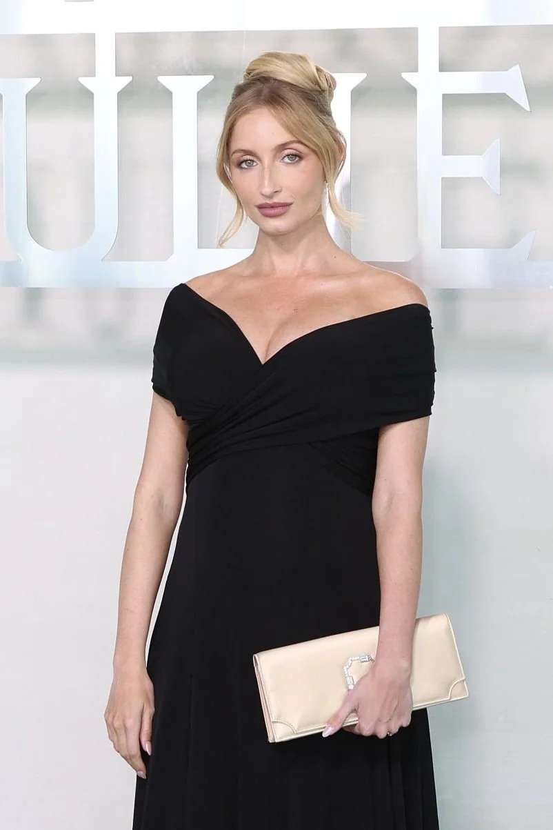 A woman with blonde hair styled in an updo, wearing a black off-the-shoulder gown, holding a beige clutch purse, standing in front of a backdrop with large silver letters.
