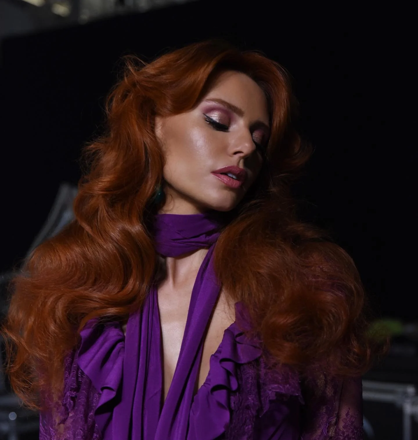 A woman with long, wavy red hair, closed eyes with pink eyeshadow, and wearing a purple dress with ruffles.