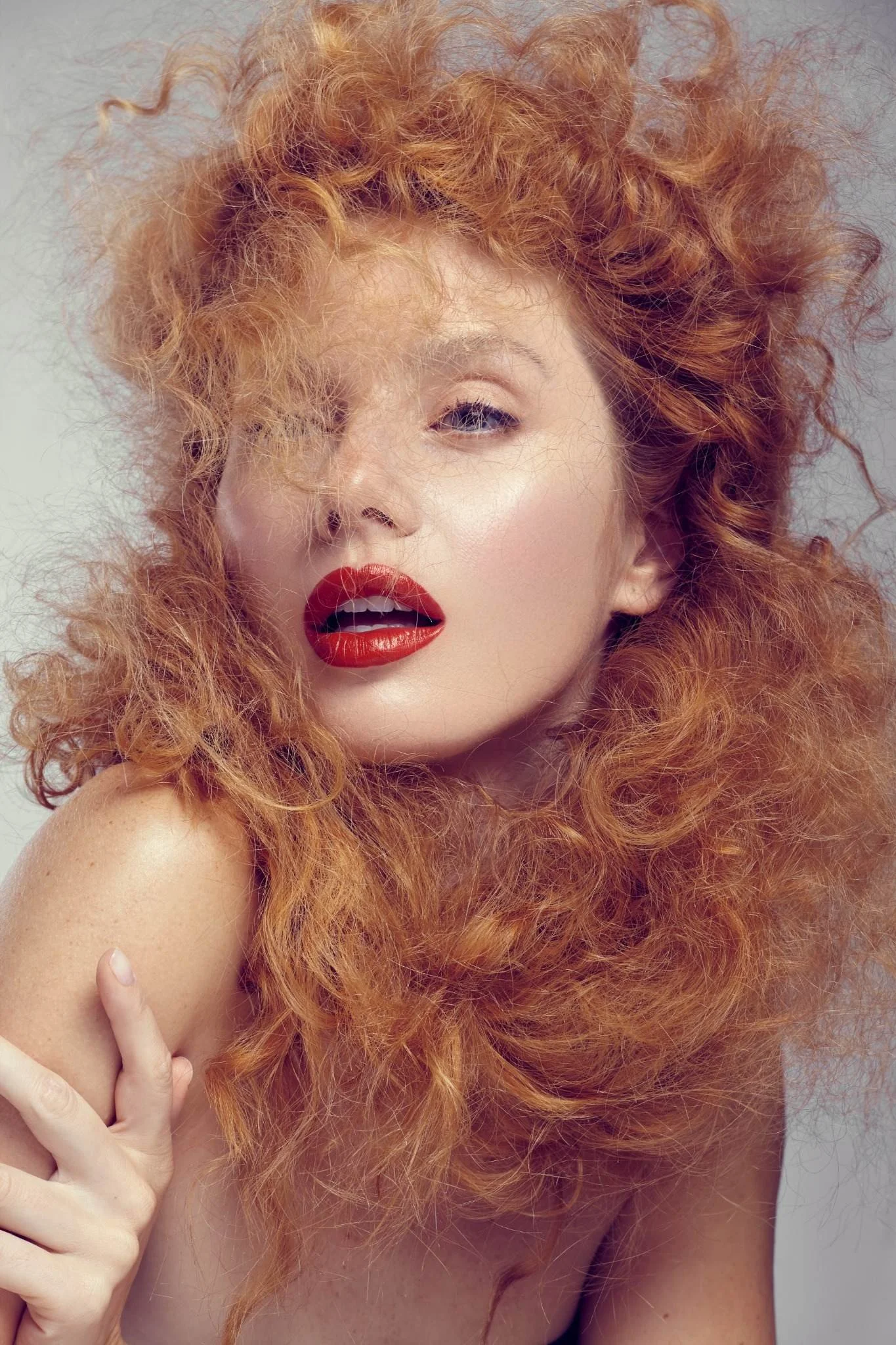A woman with long, curly red hair and red lipstick poses with her hand near her face, partially covering her eye, against a plain background.