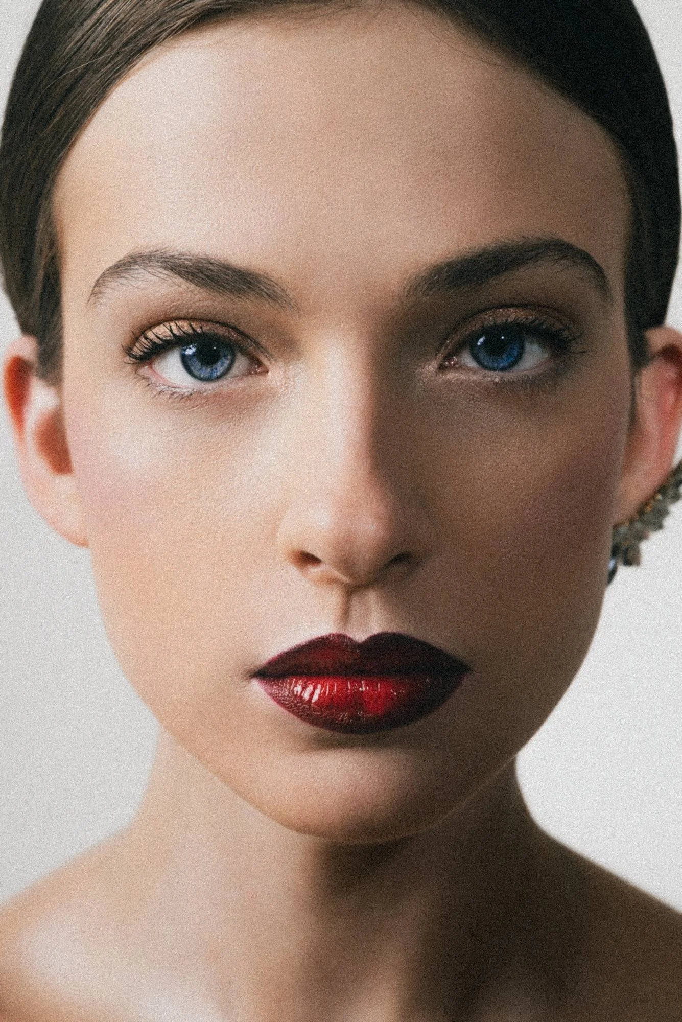 Close-up of a woman's face showing her blue eyes, dark eyebrows, wearing dark red lipstick, with dark hair and a neutral background.