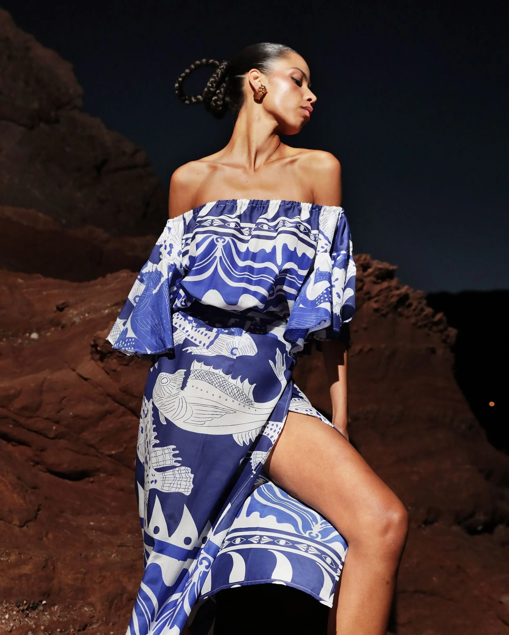 A woman with braided hair and gold earrings posing outdoors at night, wearing an off-shoulder blue and white patterned dress, with rocky terrain and a dark sky in the background.