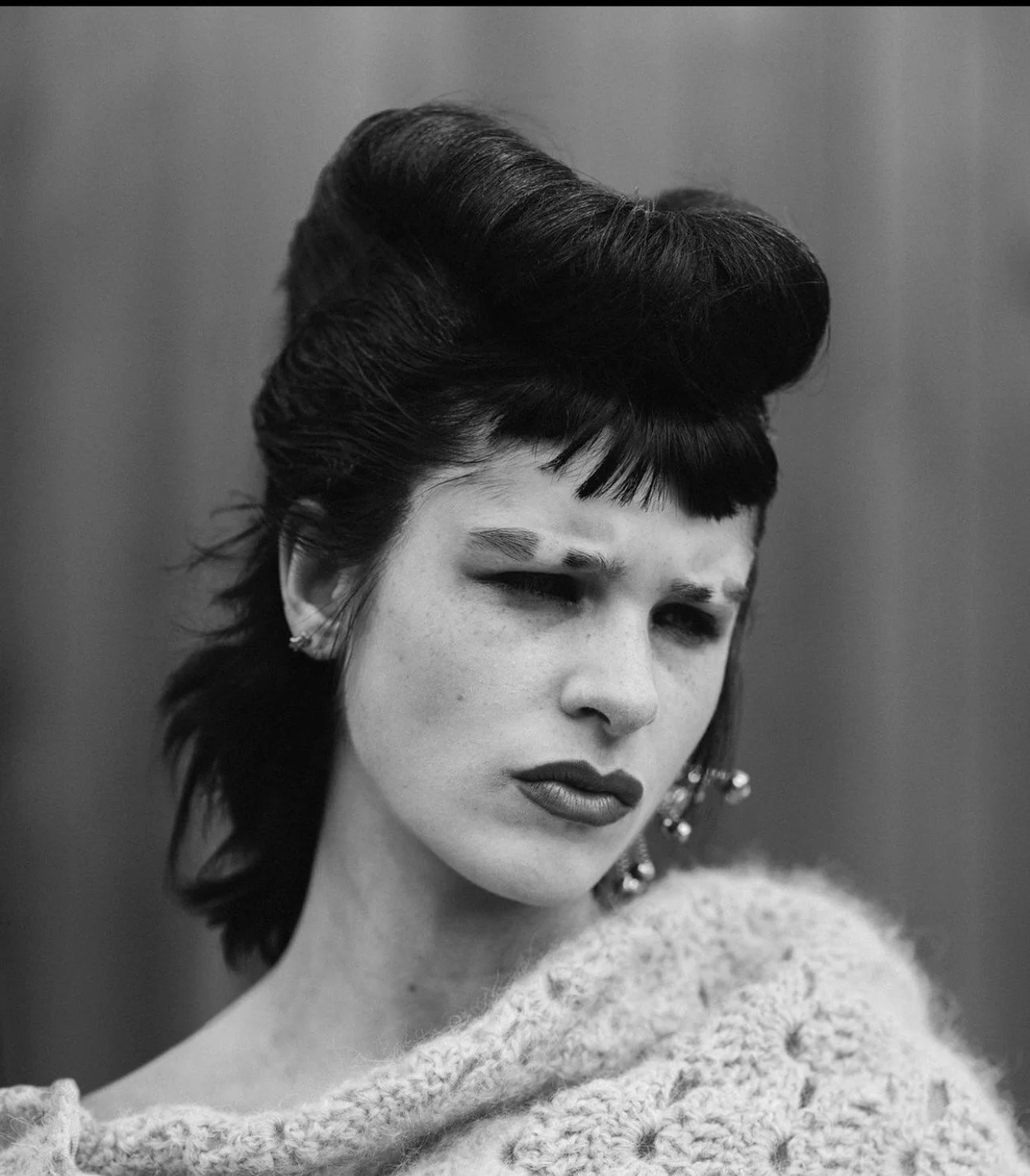 Black and white portrait of a woman with styled dark hair, bold makeup, and earrings, wearing a textured sweater.