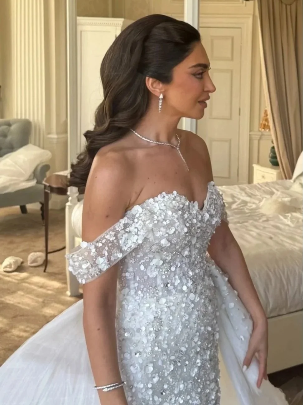 A woman dressed in a white, off-shoulder, floral-embellished gown stands inside a bedroom, with elegant curtains, a bed, and furniture visible in the background.