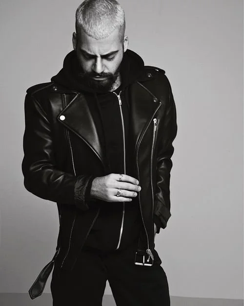 A man with short, light-colored hair and a beard, wearing a black leather jacket over a hoodie, looking down with his hand touching his jacket, against a plain background.