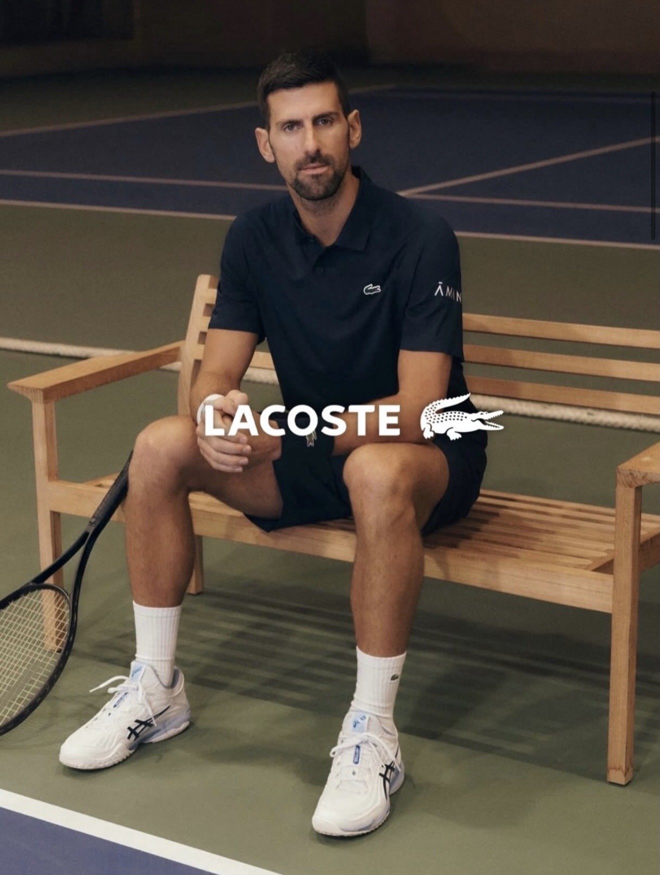 A man sitting on a wooden bench on a tennis court, holding a tennis racket, wearing a black Lacoste polo shirt, black shorts, white Nike sneakers, and white socks. The Lacoste logo is visible on his shirt and shoes, and the Lacoste logo and name are 