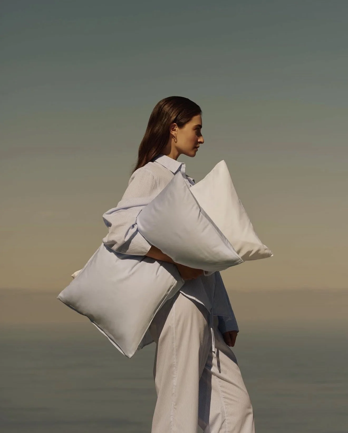 A woman holding pillows stands against a gradient sky background, with her head side profile facing right.
