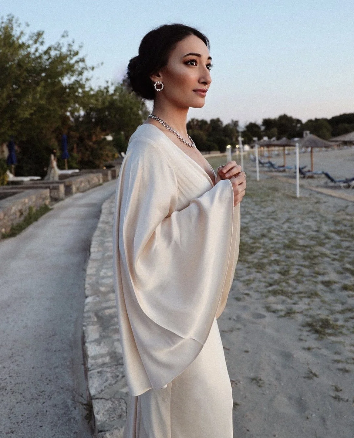 A woman dressed in an elegant cream-colored gown standing on a beachside pathway during dusk, with her hands clasped near her chest, wearing jewelry including earrings and a necklace.