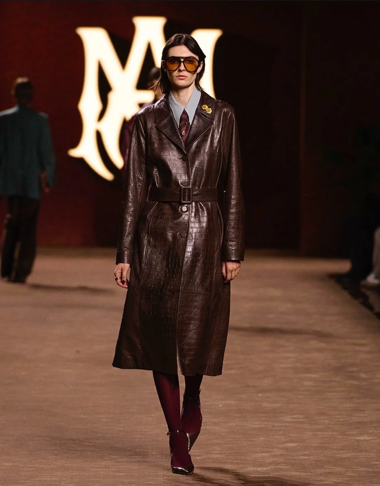A female model walking down a runway wearing a long brown leather trench coat with a belt, maroon stockings, and black shoes with heels. She has long dark hair, large tinted sunglasses, and a small yellow pin on her coat. A large illuminated logo is 
