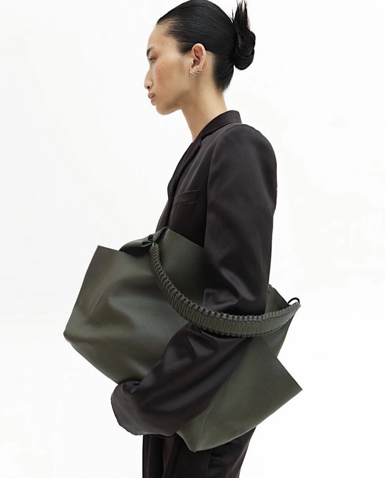Side profile of a woman with dark hair in a bun, wearing a black blazer, carrying a large olive-green bag with a ridged handle, standing against a plain white background.