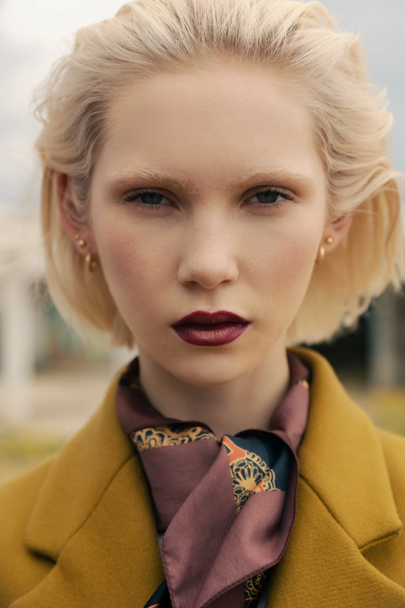 Close-up of a woman with platinum blonde hair, blue eyes, wearing dark lipstick, earrings, a mustard yellow coat, and a patterned silk scarf.
