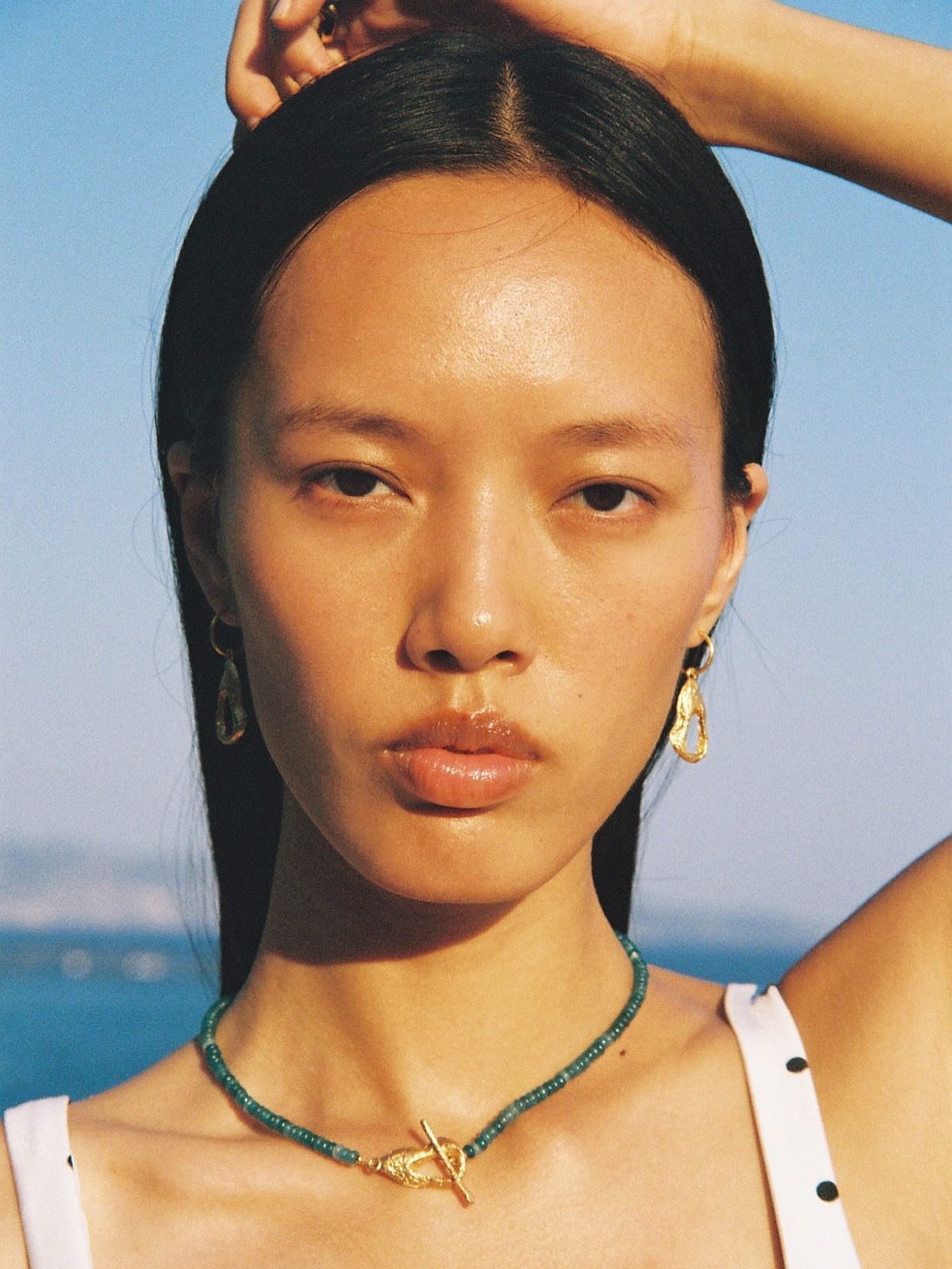 A young woman with long black hair, wearing gold earrings and a necklace, holding her arm above her head, with a blue sky and ocean in the background.