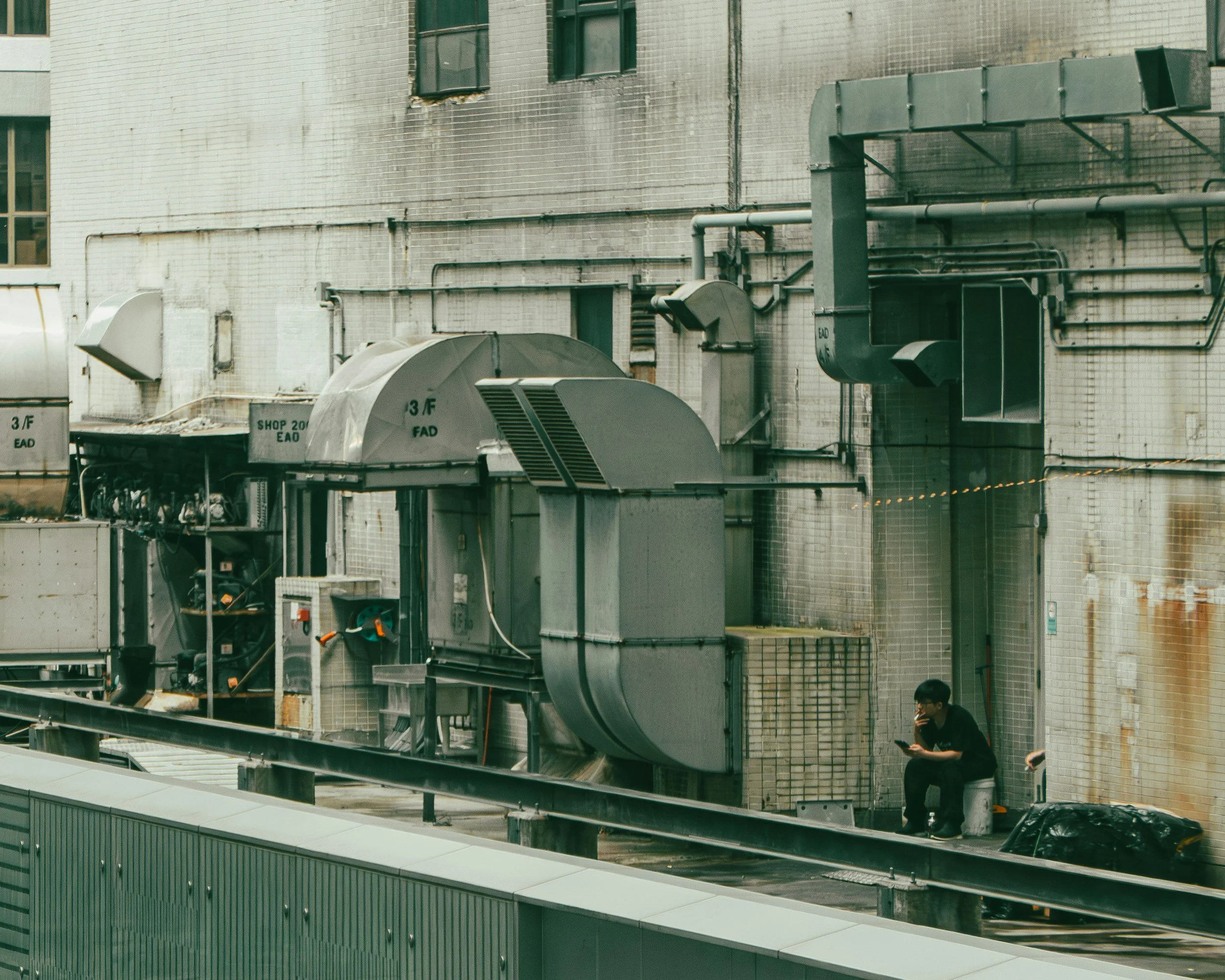 An urban building wall with multiple HVAC units and ventilation pipes, with two people at the bottom, one walking and one talking on a phone.