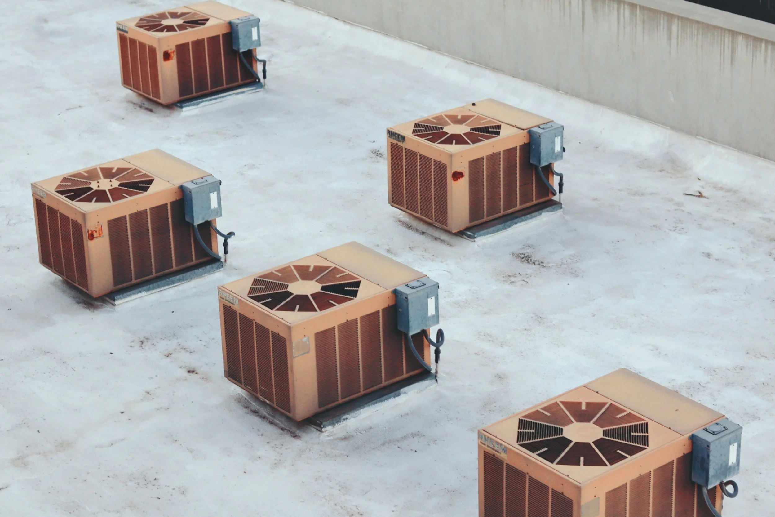Multiple rooftop air conditioning units installed on a flat rooftop with a concrete wall in the background.