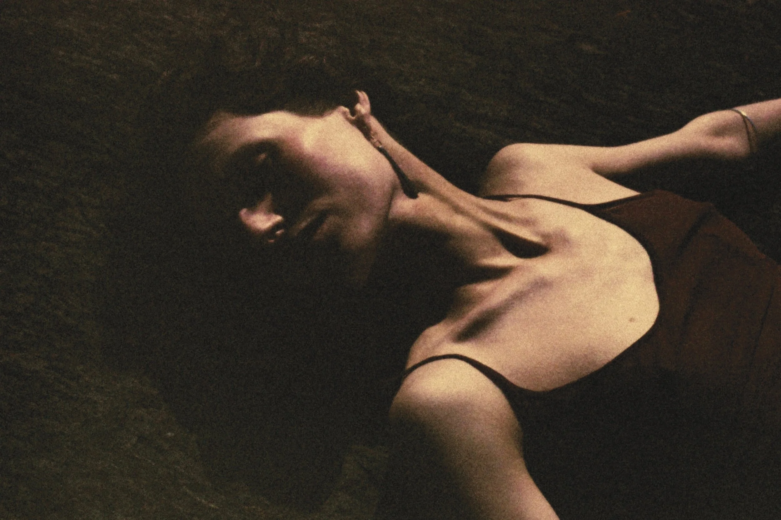 A woman with short dark hair lying on a dark surface, wearing a black sleeveless top, with her eyes closed and her hand near her face.