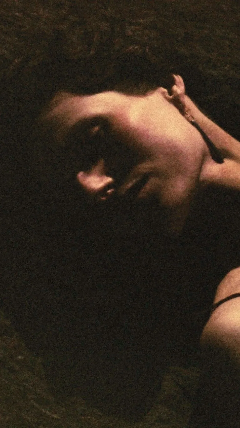 A person with short dark hair lying down in a dark setting, smiling, with their hand resting on the side of their face.