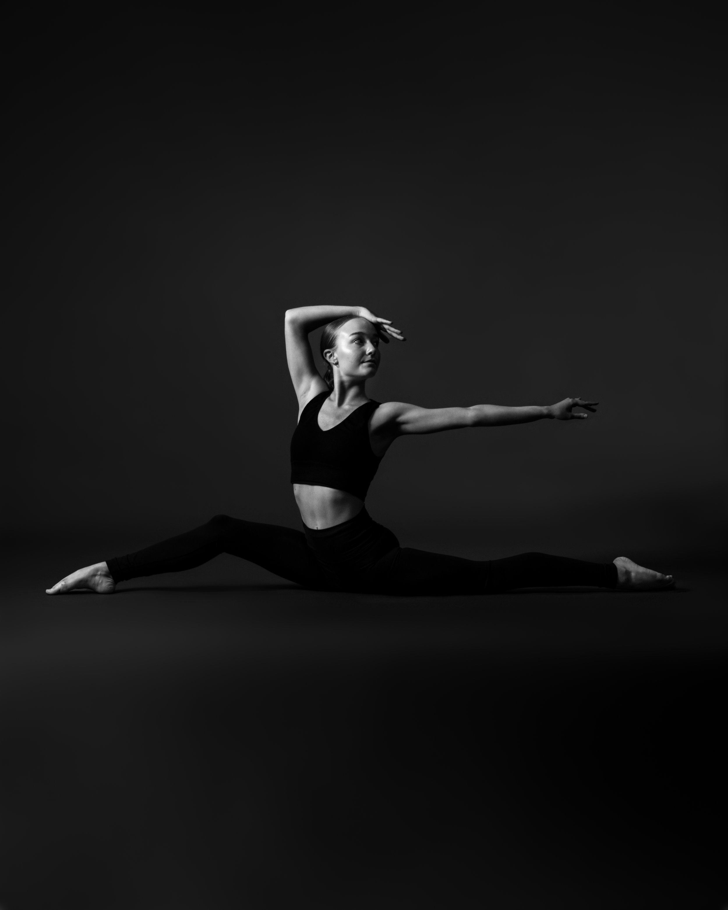 Black and white photo of a woman performing a dance pose on a dark background, with her right arm raised above her head and her left arm extended outward, legs in a split position, wearing a black sports bra and black leggings.