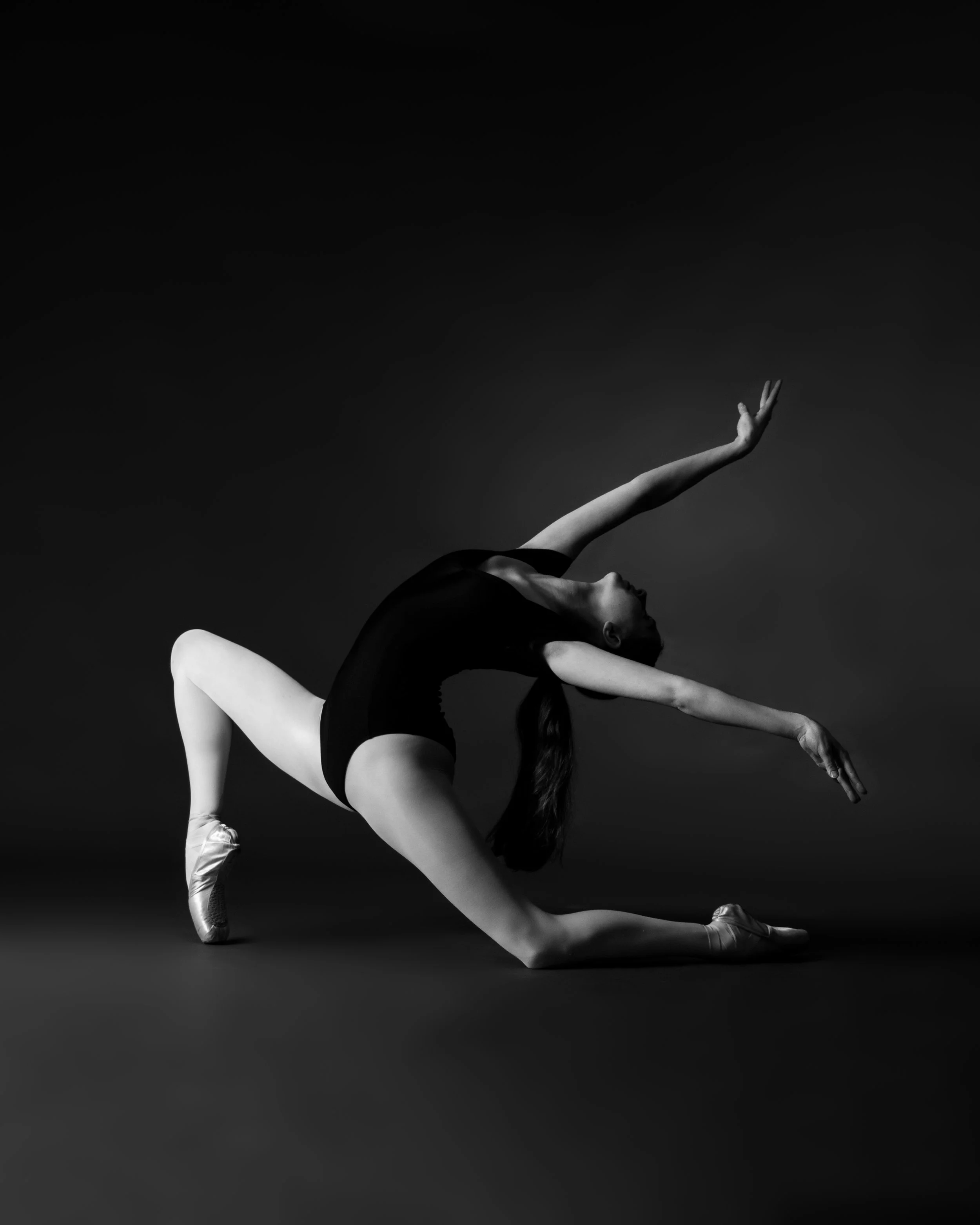A black and white photo of a ballet dancer performing a pose with one arm raised and the other reaching downward, wearing a leotard and ballet shoes.