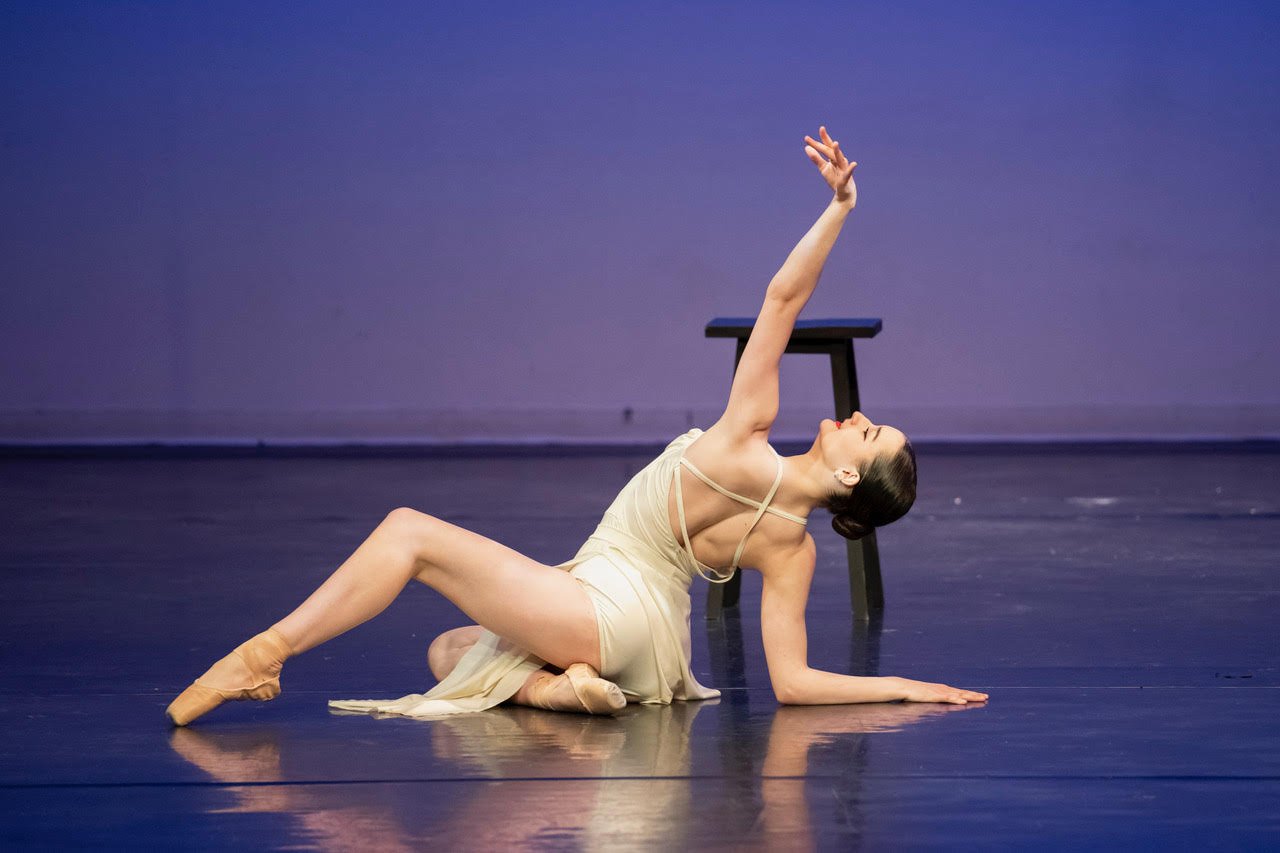 Ballet dancer on stage, posing with one arm raised and the other on the floor, wearing a beige dress and pointe shoes, with a chair in the background.