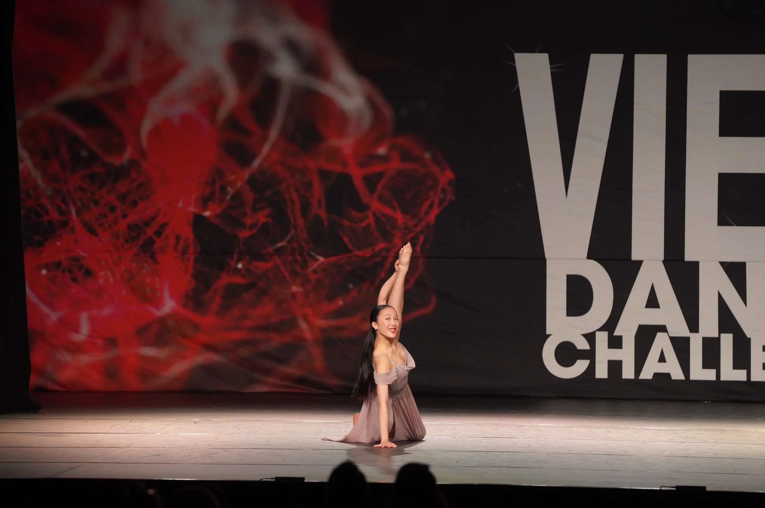 A woman performing a dance move on stage, kneeling on one knee with one arm extended upward, with a mostly red and black background and a large text display that reads "THE VIE" and some additional words partially visible.