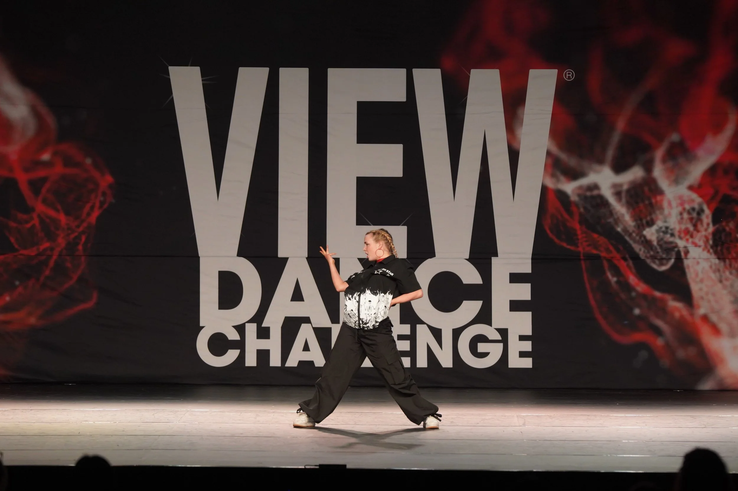 A young woman dancing on stage in front of a large backdrop that reads 'VIEW' and 'CHALLENGE' with red patterns.