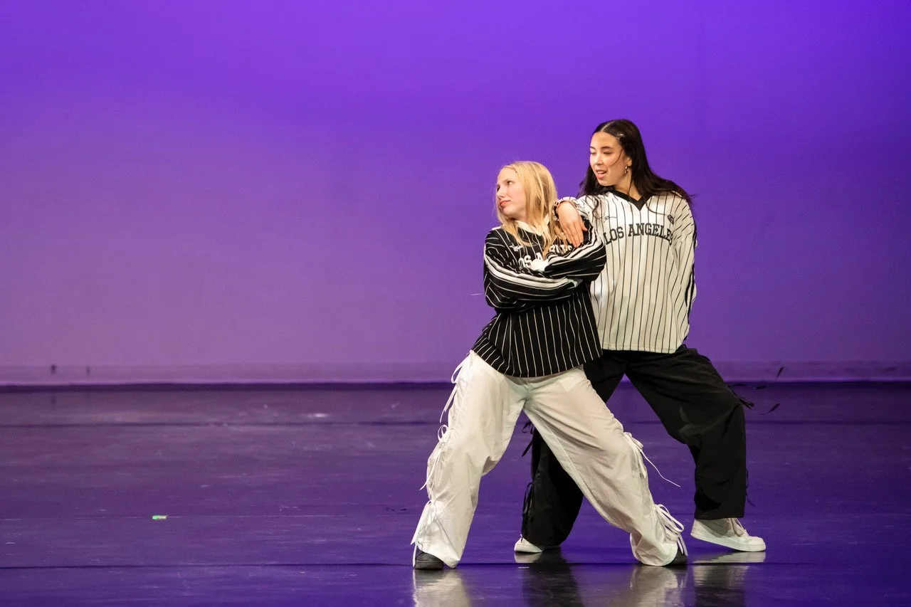 Two women posing on stage with purple backdrop, wearing casual streetwear, one with striped shirt and white pants, the other with oversized baseball shirt and black pants.