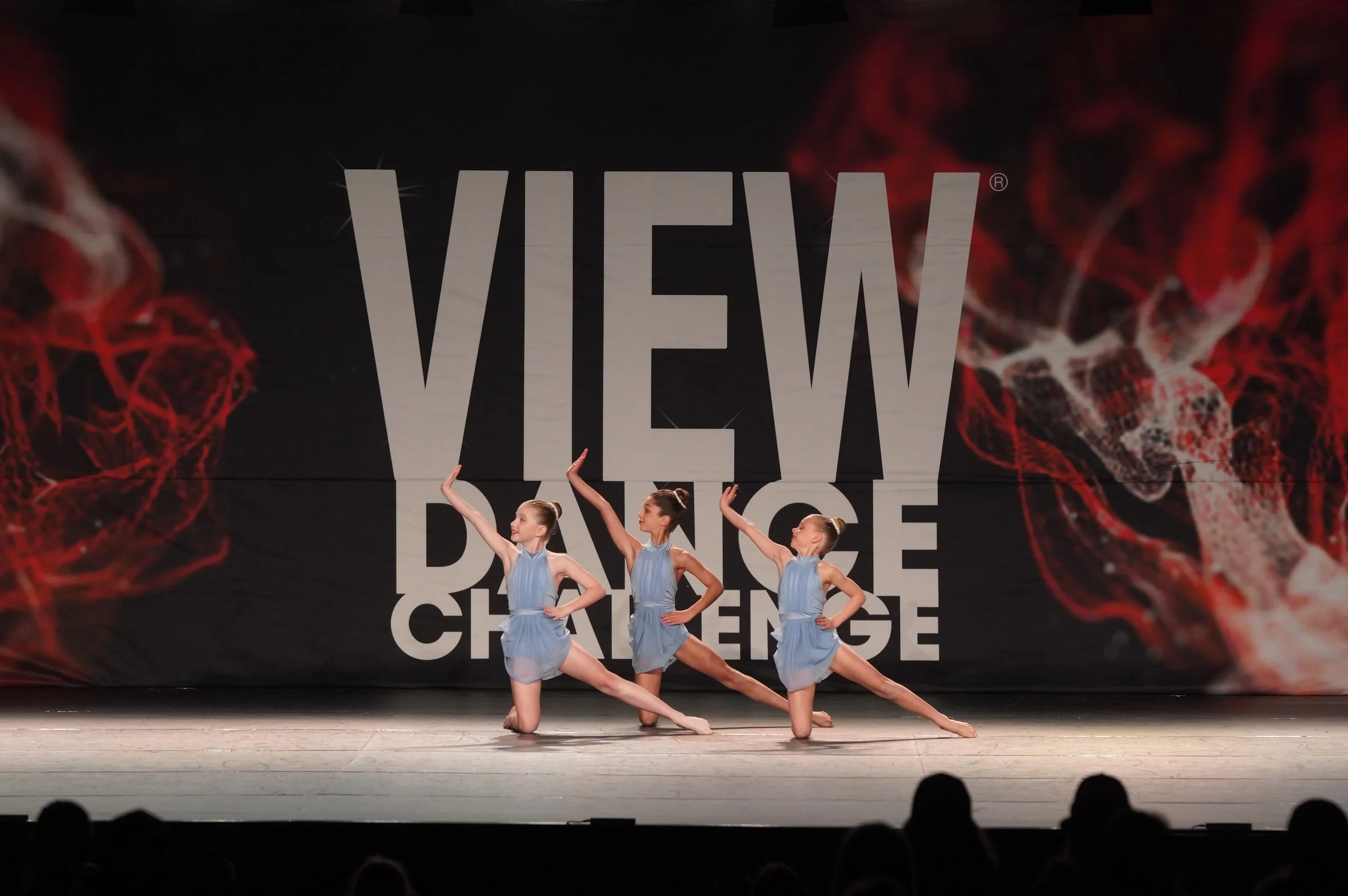 Four young ballet dancers in blue costumes perform a dance routine on stage in front of a large sign that reads 'VIEW', with a red and black background design.