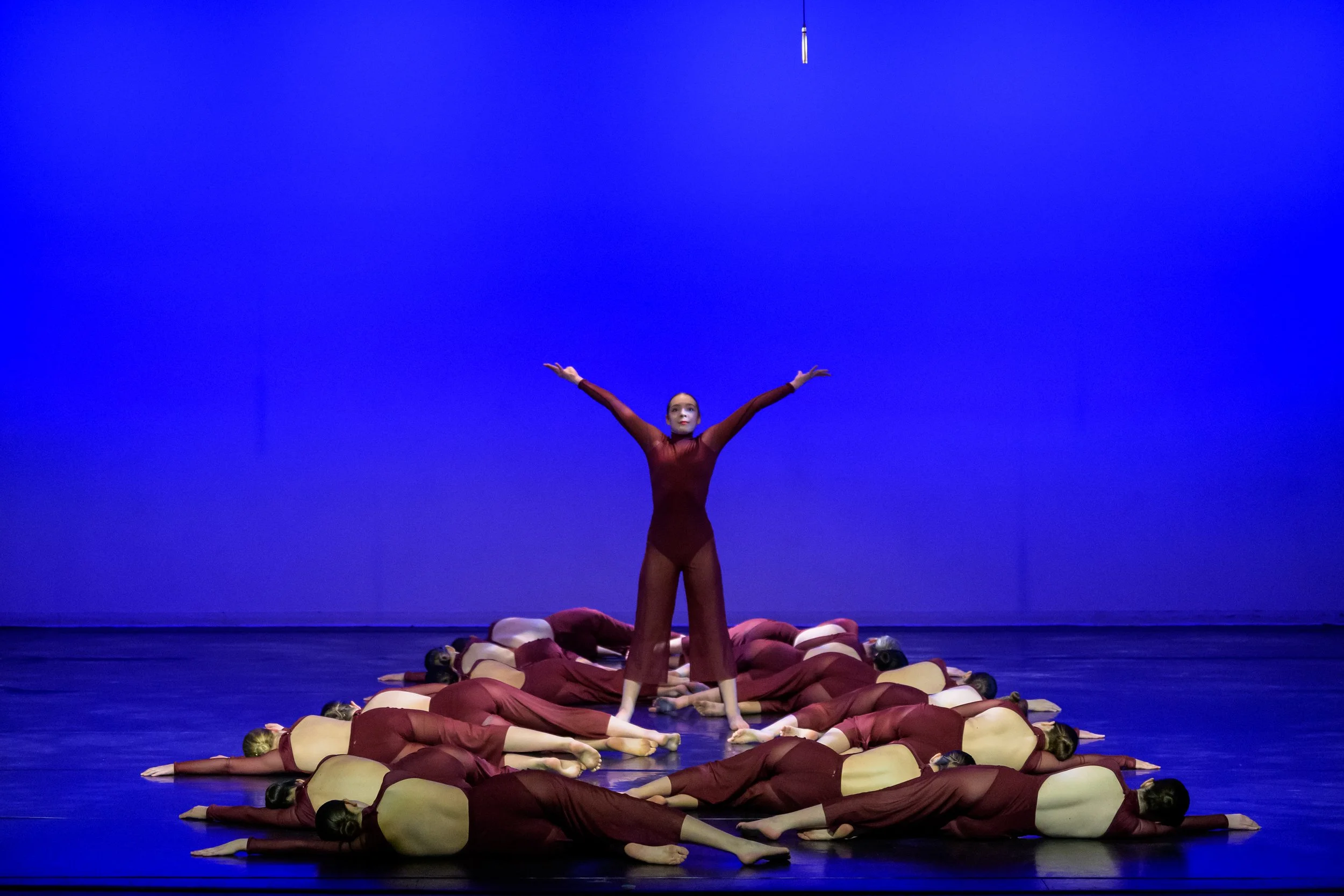 A group of dancers is performing on a stage with a blue backdrop. One dancer is standing with arms outstretched, while others are lying face down on the floor with arms by their sides, dressed in matching maroon costumes.