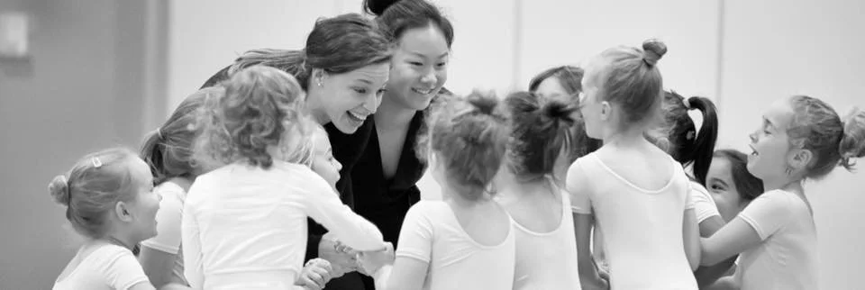 A group of young girls and two women in a dance studio, smiling and interacting.