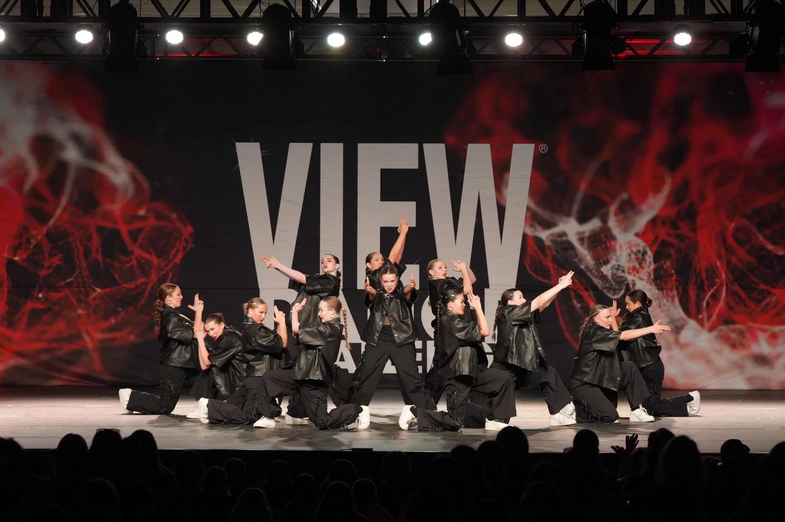 A group of young women dancers performing on a stage with a background displaying the words "VIEW" and red, smoky graphics.