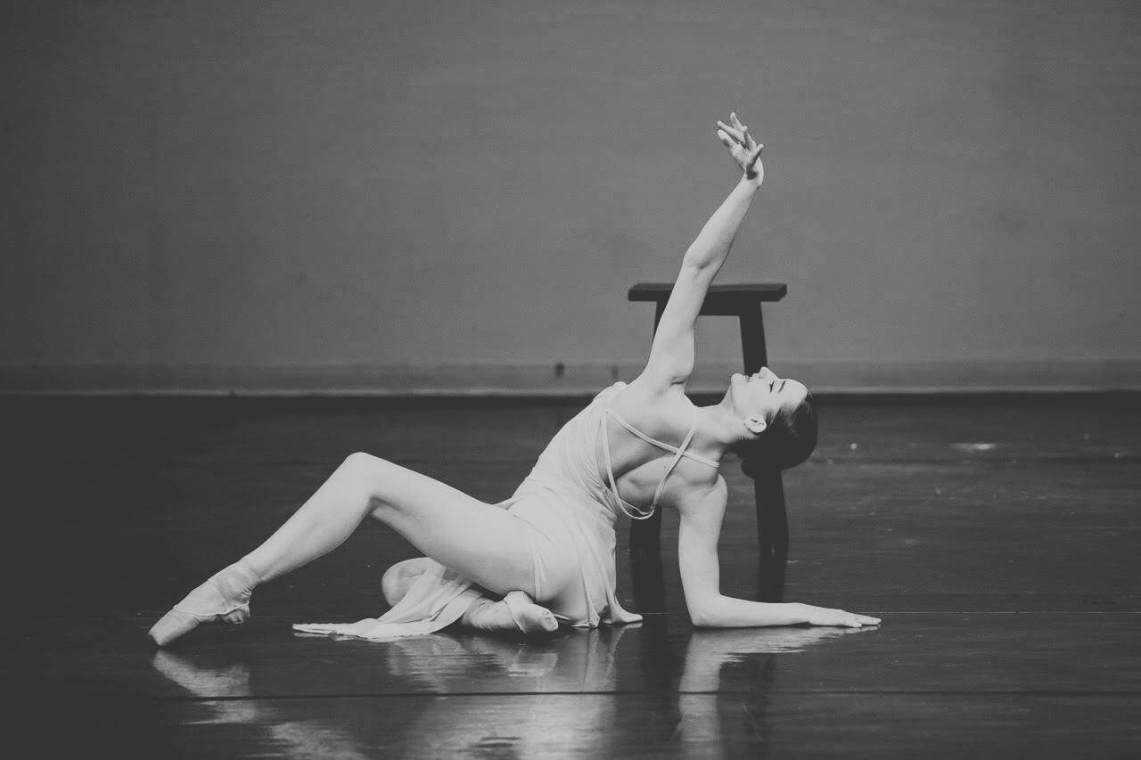 A female ballet dancer practicing on stage, kneeling on the floor with one hand on the ground and the other arm extended upwards, wearing a light-colored ballet costume and pointe shoes, with a chair behind her.