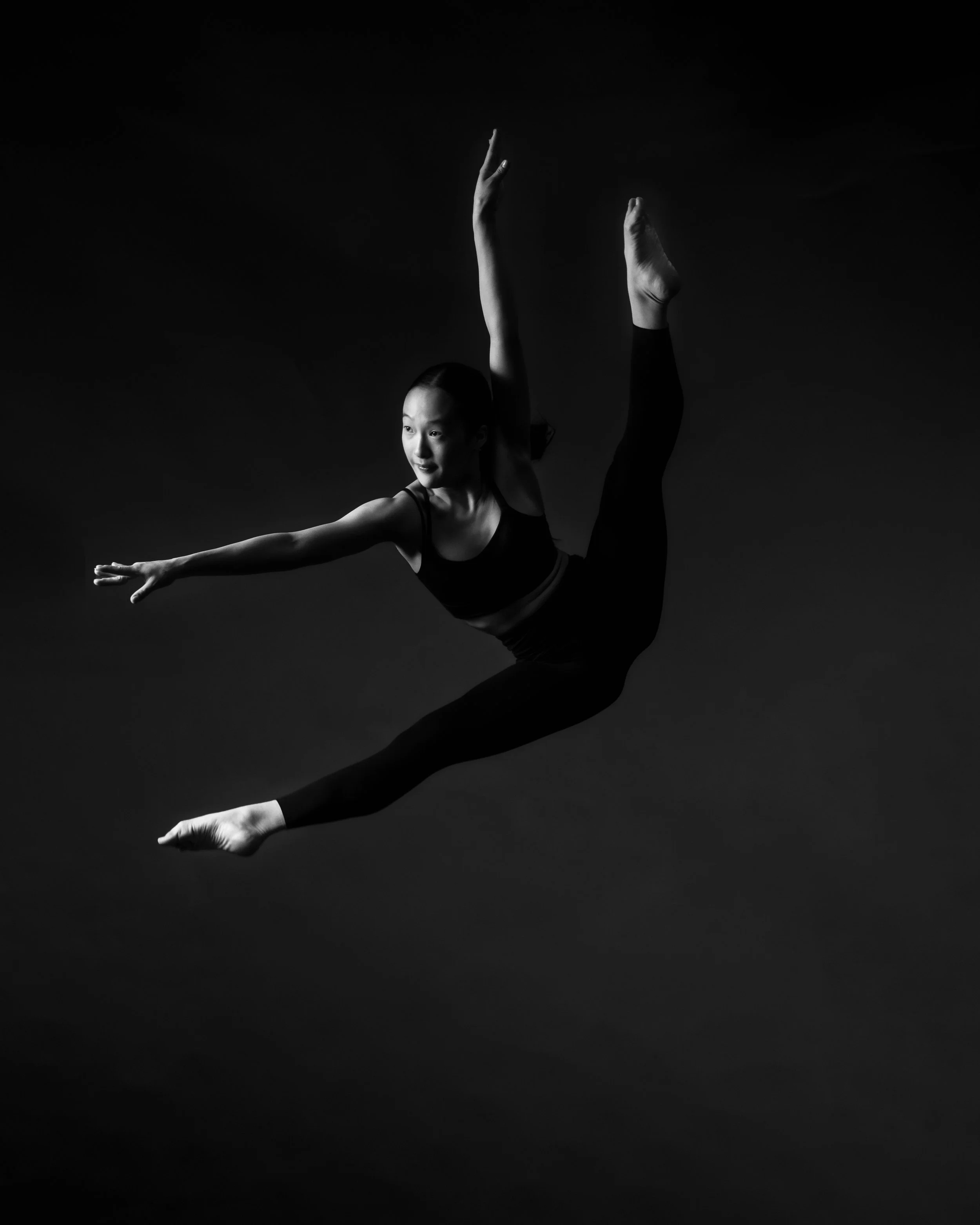 A black and white image of a female ballet dancer performing a pose with one leg extended high and arms outstretched, against a dark background.