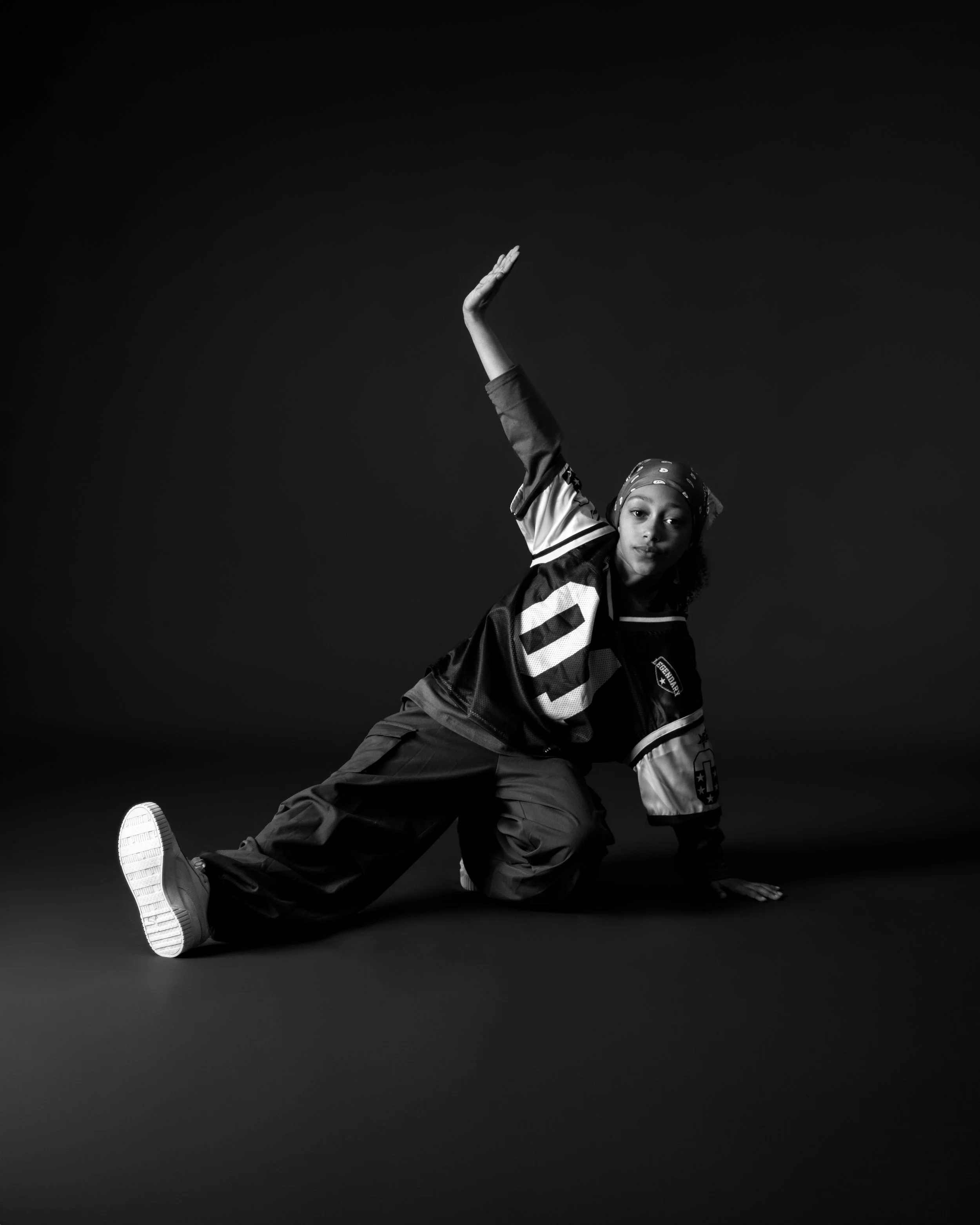 Black and white photo of a young woman wearing a bandana and sports jersey, performing a dance move on a dark background.