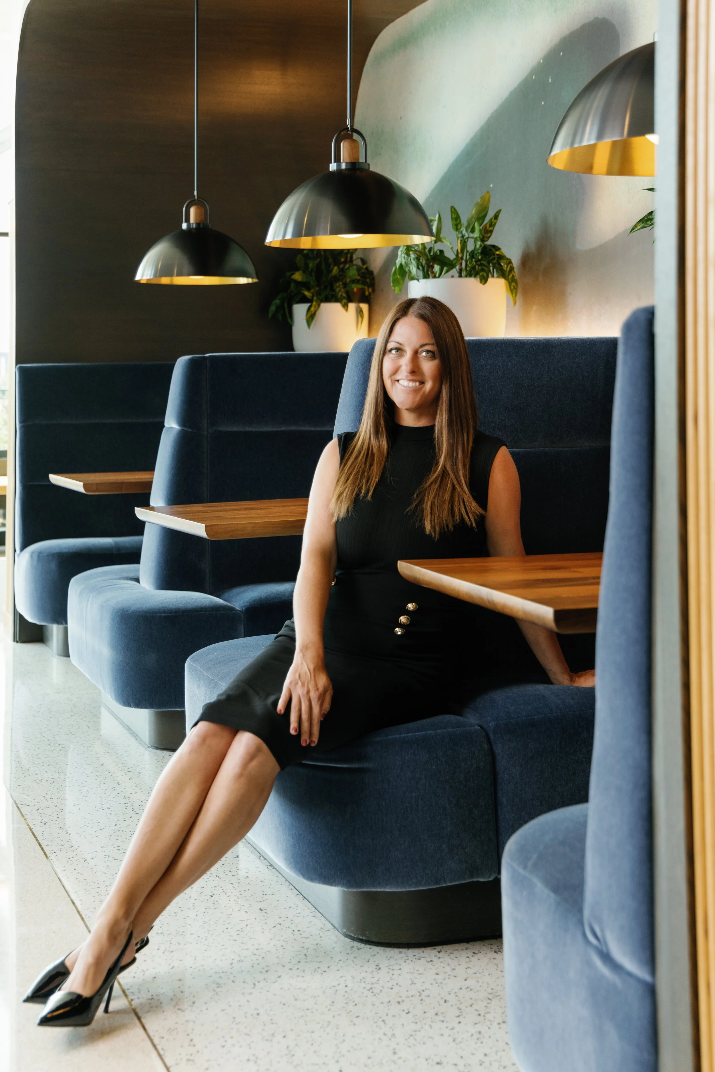 A woman in a black sleeveless dress and high heels sitting in a booth at a restaurant or cafe, smiling at the camera. The booth has blue upholstered seats, wooden tables, and the background includes hanging black pendant lights and large potted plants against a painted wall.