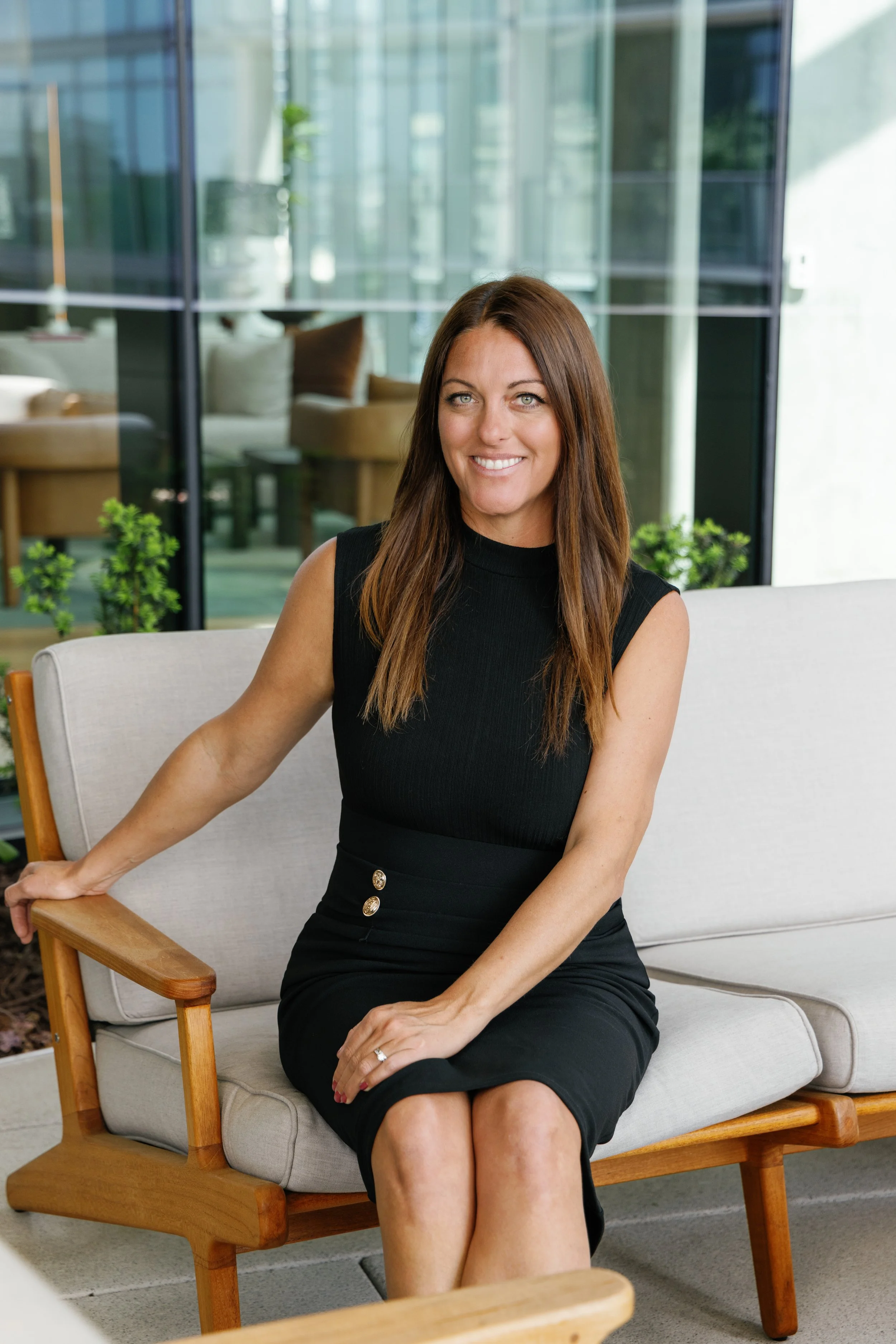 A woman with long brown hair in a black sleeveless dress sitting on a light-colored sofa with a wooden frame and armrest, smiling, in a modern indoor space with large windows and green plants.