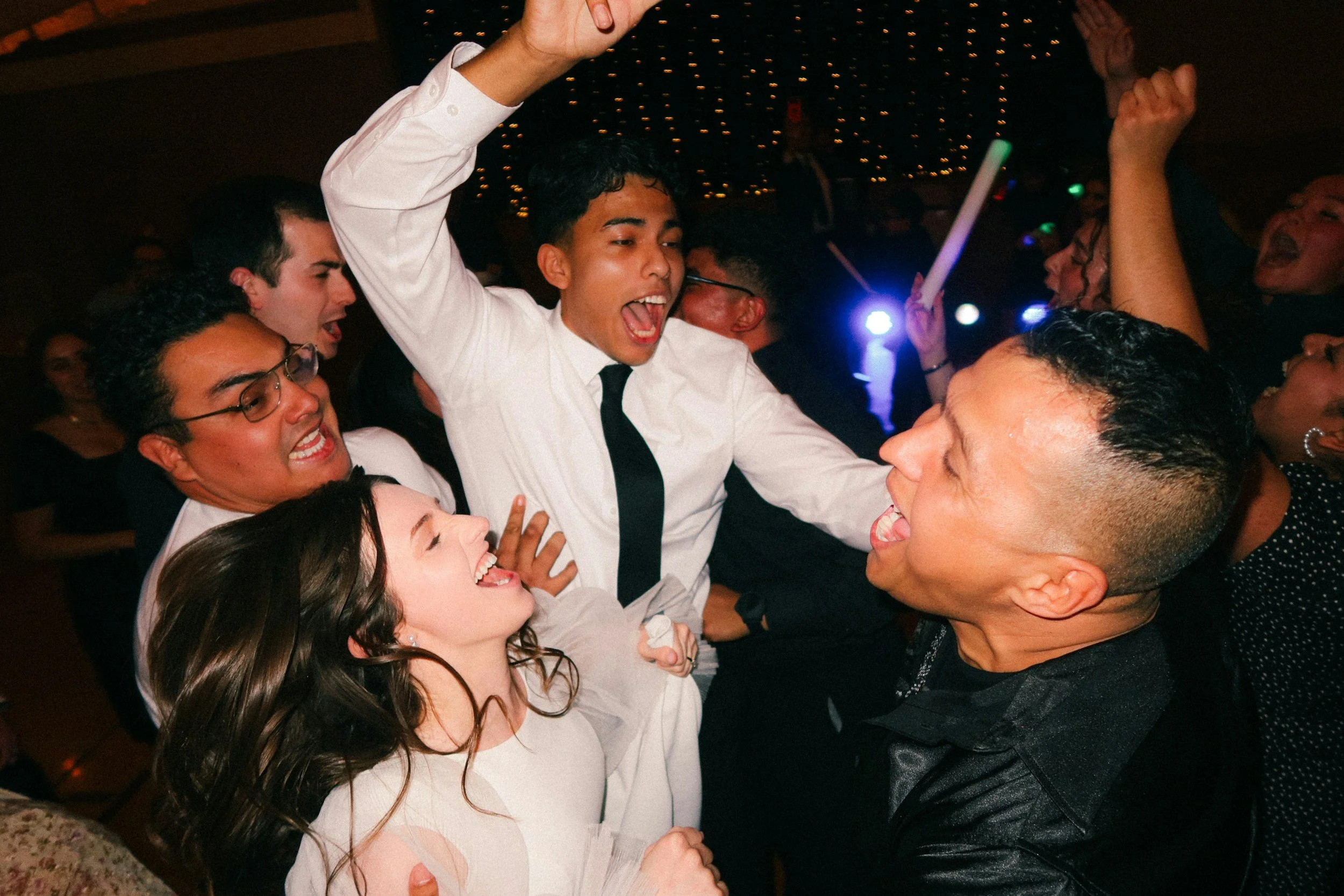 A group of young people at a party, singing and dancing together. The scene is lively and festive, with colorful lights in the background. The central young man is wearing a white shirt and black tie, with his arms raised and mouth open, enjoying the moment.