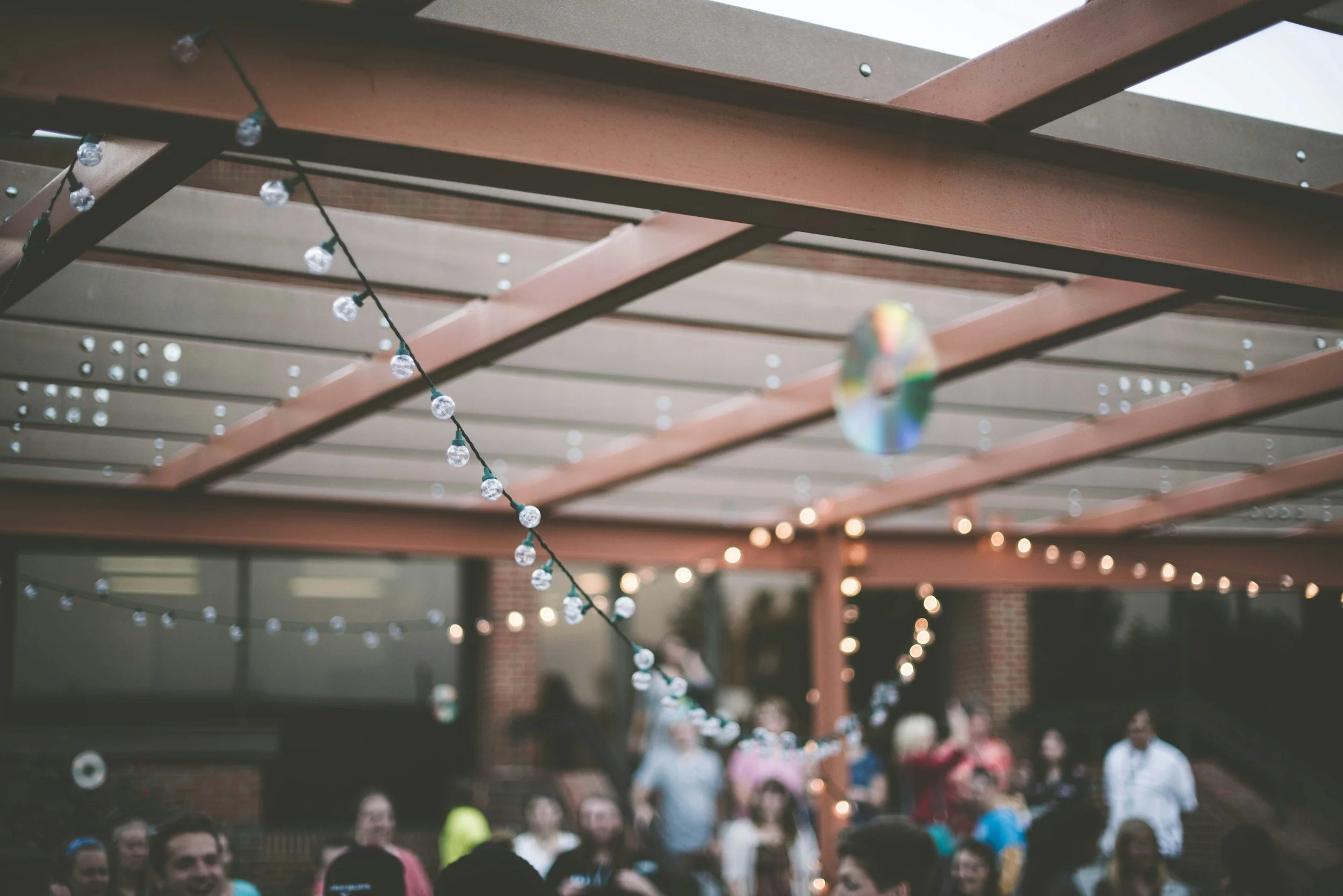String of decorative string lights hanging under a wooden pergola with people in the background at an outdoor gathering.