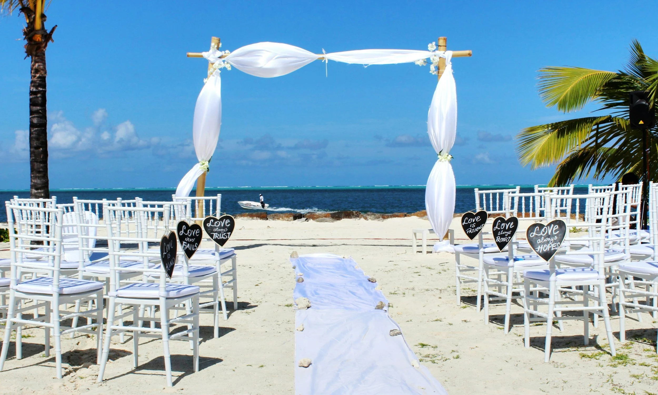 A beach wedding setup with white chairs arranged on sand, decorated with black heart-shaped signs with white inscriptions, a white aisle runner, and a bamboo wedding arch draped with white fabric. The ocean and blue sky are visible in the background, with palm trees on either side of the setup.
